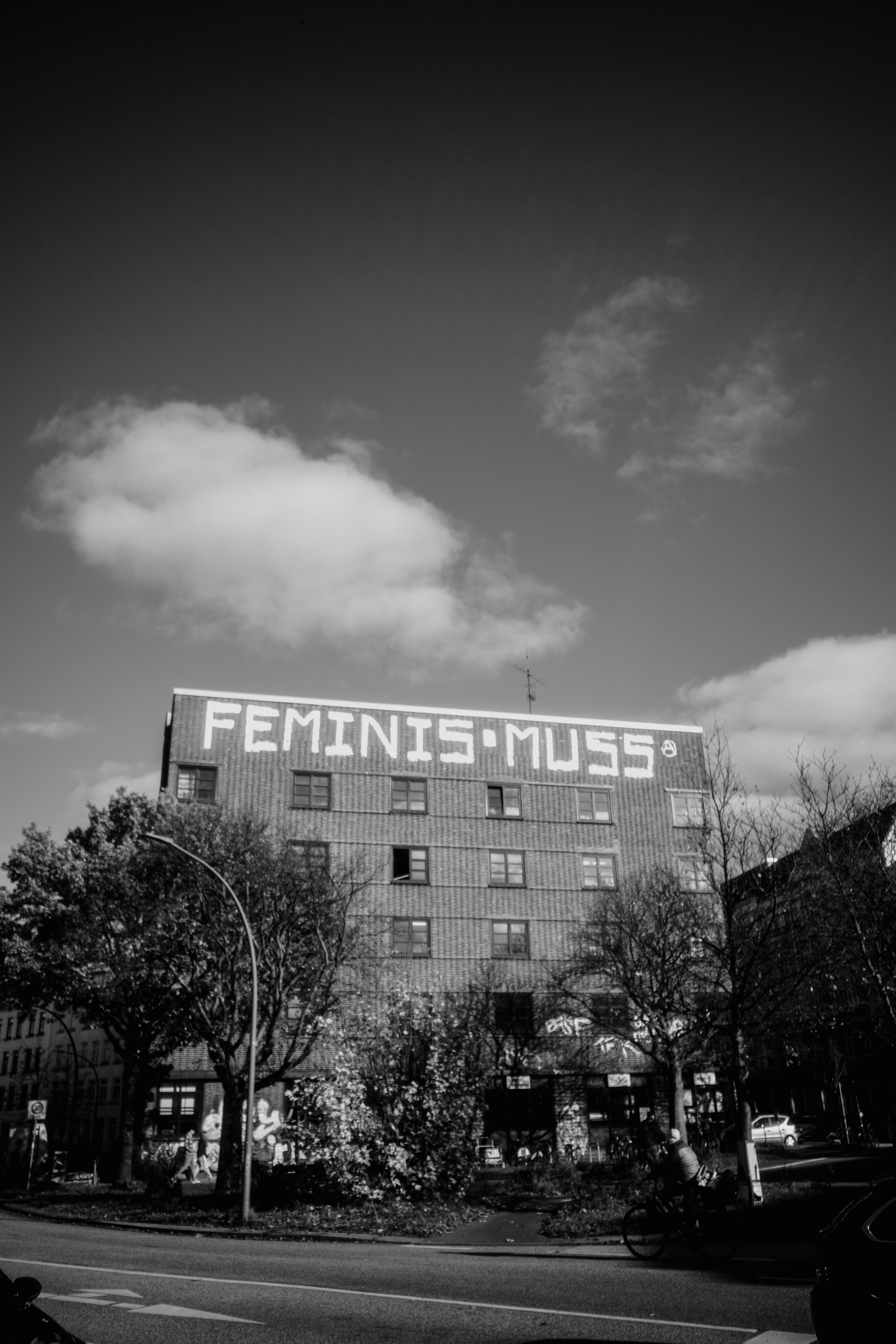 The image shows a black-and-white photograph of a multi-story building with large graffiti reading “FEMINISMUS” on the top, set against a cloudy sky with trees and a street in the foreground.