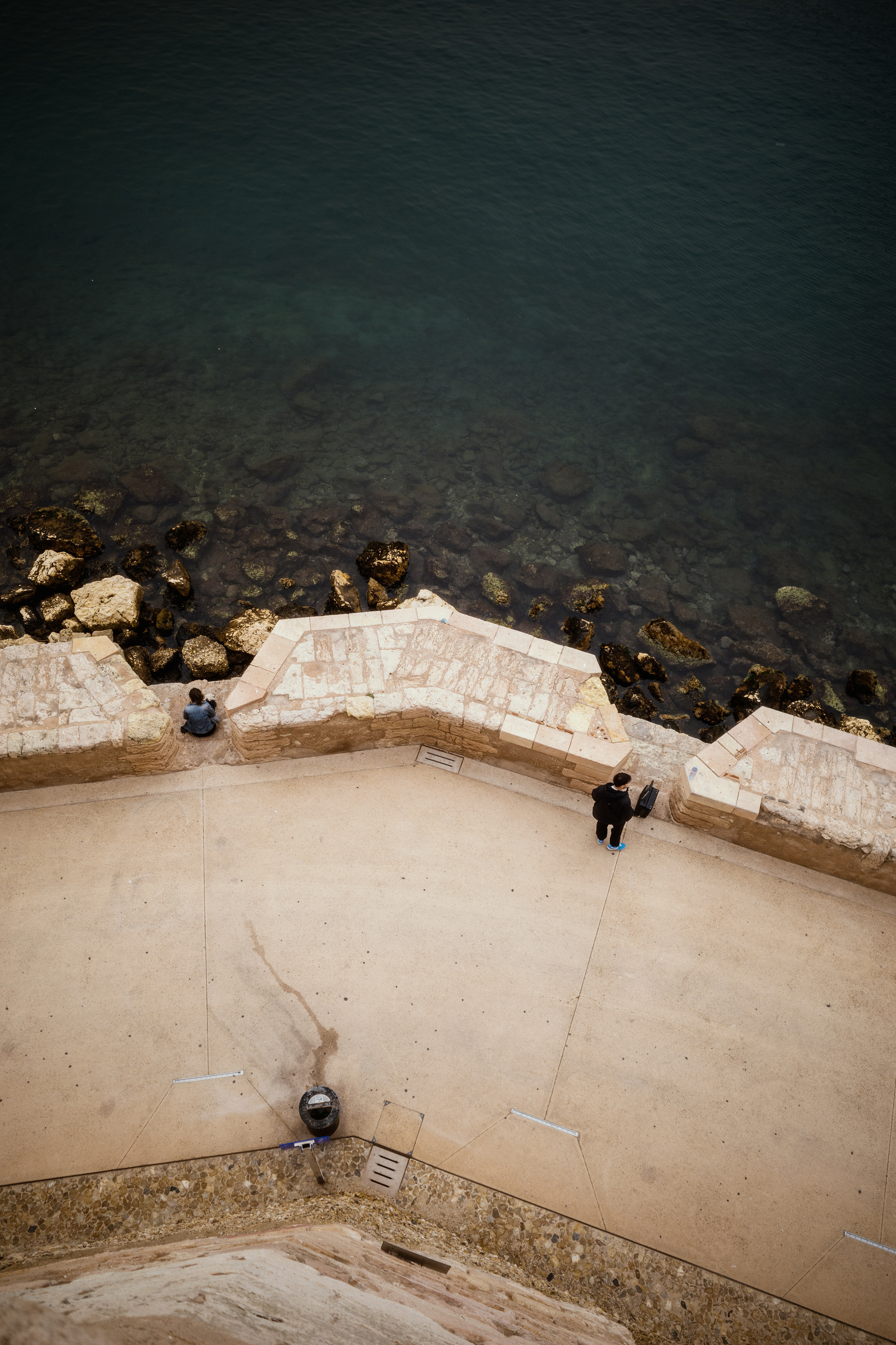 The image shows an aerial view of a stone walkway by the sea, with two people standing separately near the edge, overlooking the clear, rocky water below.