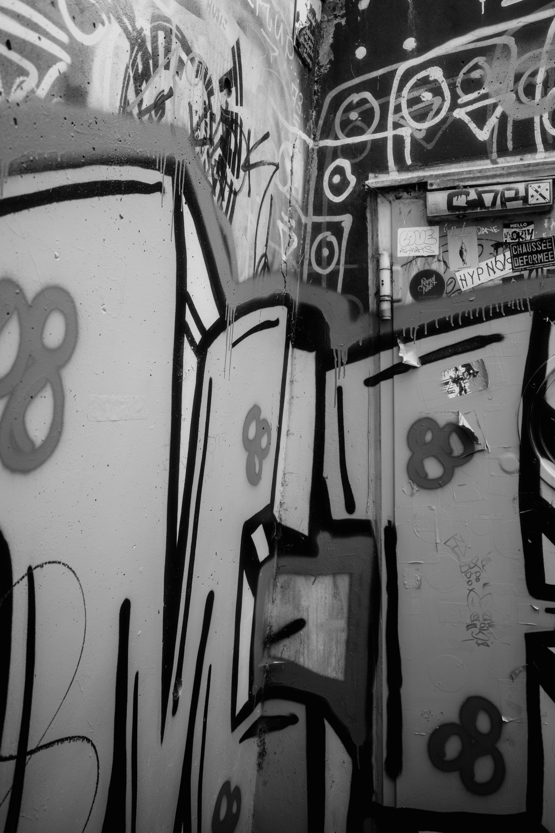 Black and white photo of a wall covered with various graffiti tags and stickers.