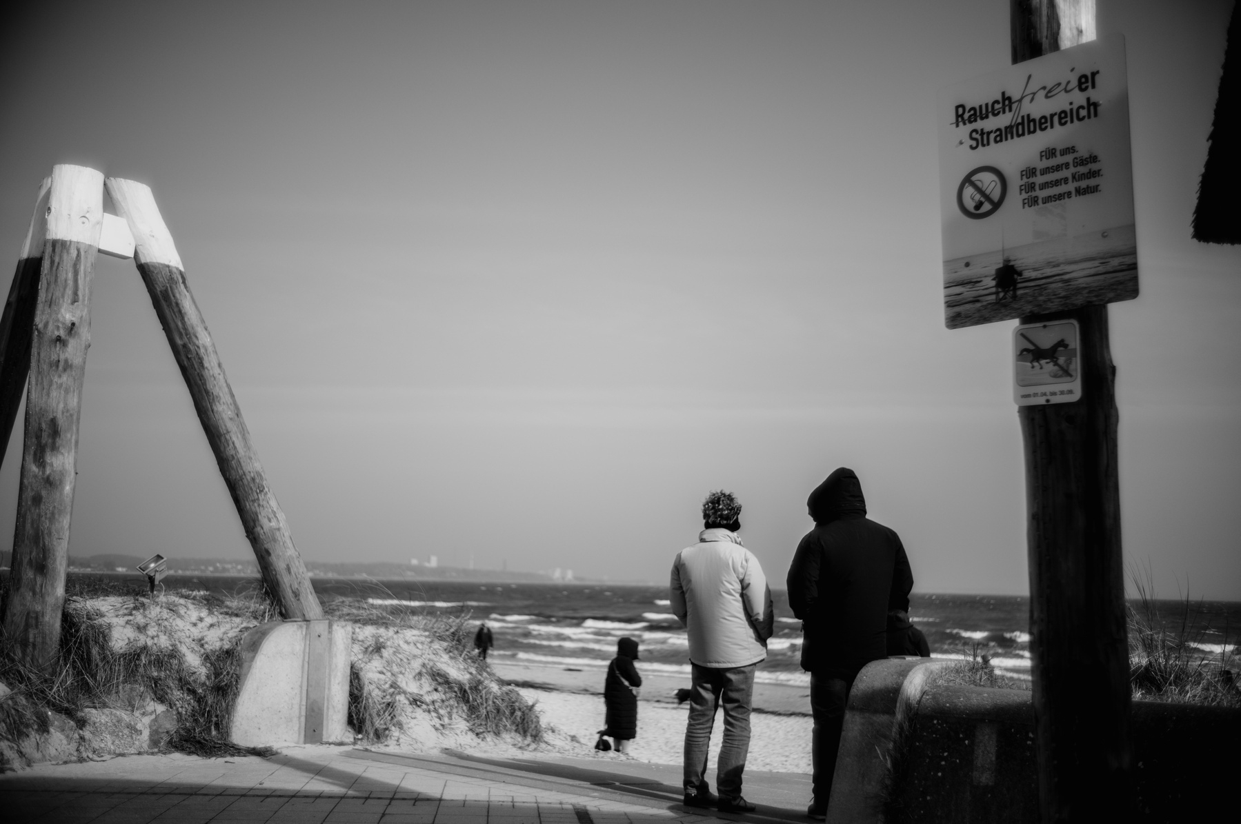 The image shows a black and white scene of two adults observing another person by the ocean on a smoke-free beach, with a clear sky and distant structures on the horizon.