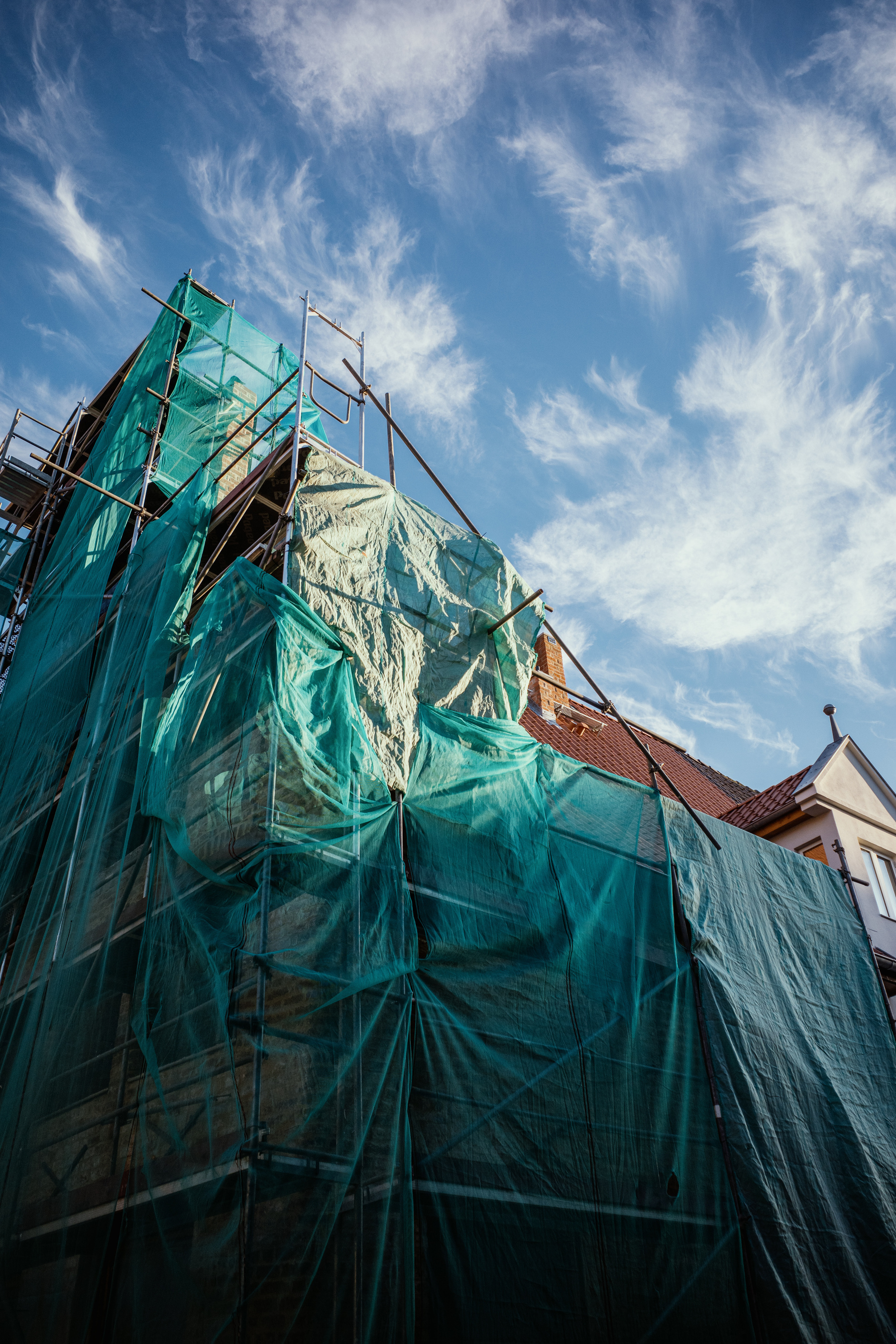 The image shows a building under construction or renovation, covered in green scaffolding nets against a backdrop of a bright blue sky with wispy clouds.