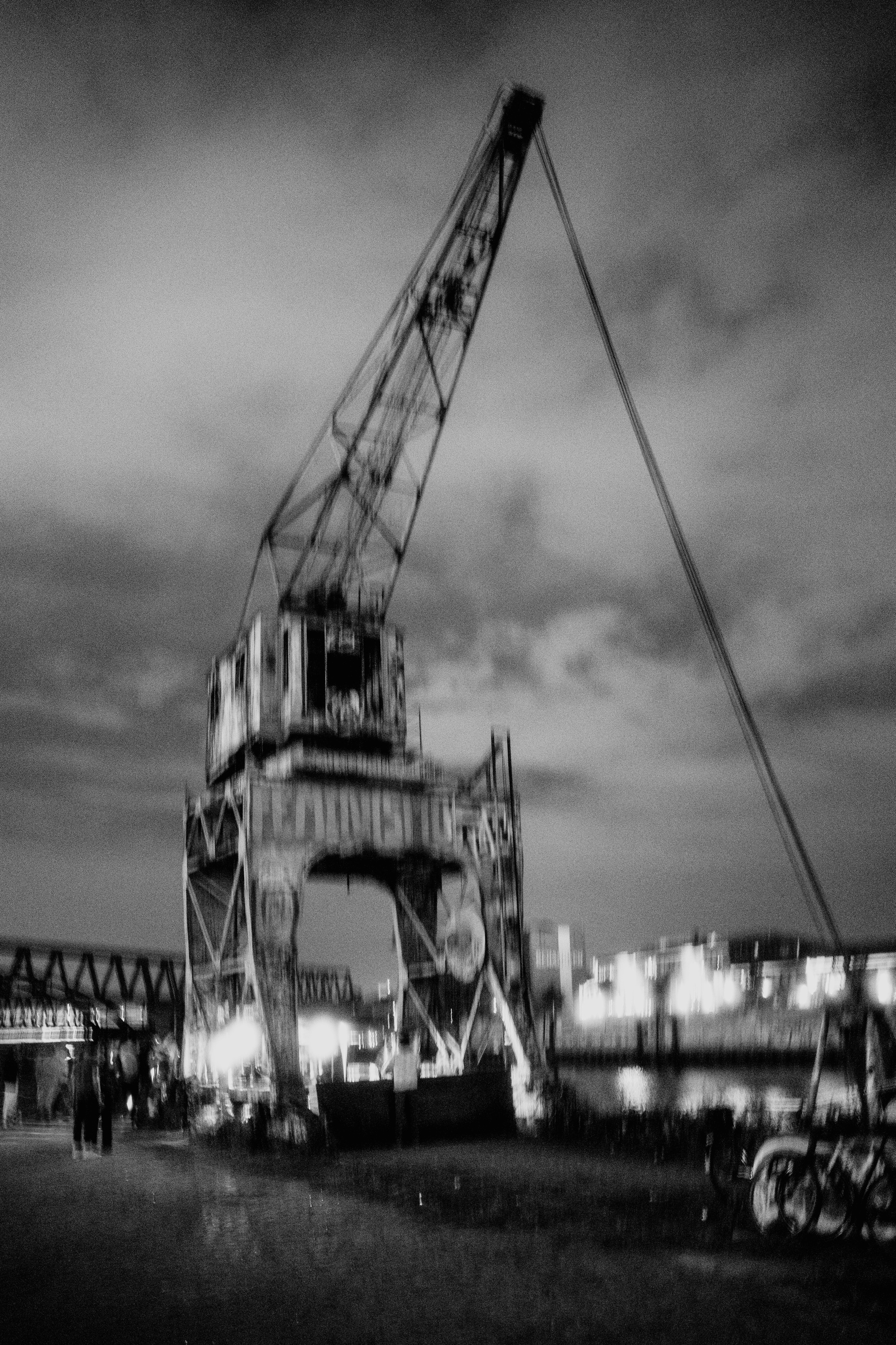 A blurry, black-and-white photo of an old, rusted industrial crane at night, with a few people and bicycles visible near the base, set against a backdrop of a cityscape and a body of water.