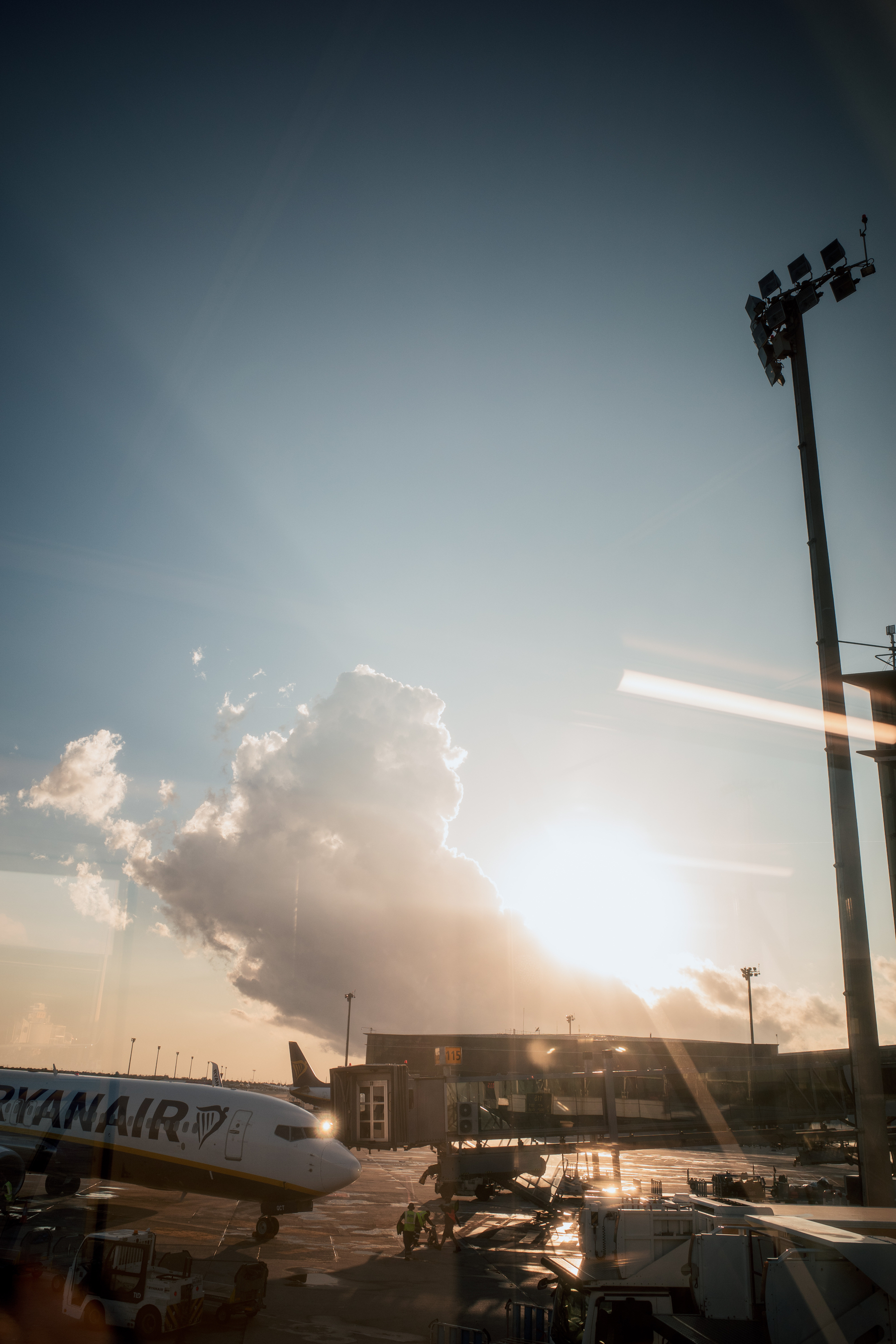 The image shows an airport runway with a Ryanair plane parked near a terminal, while the sun sets behind a large cloud, casting a warm glow over the scene.