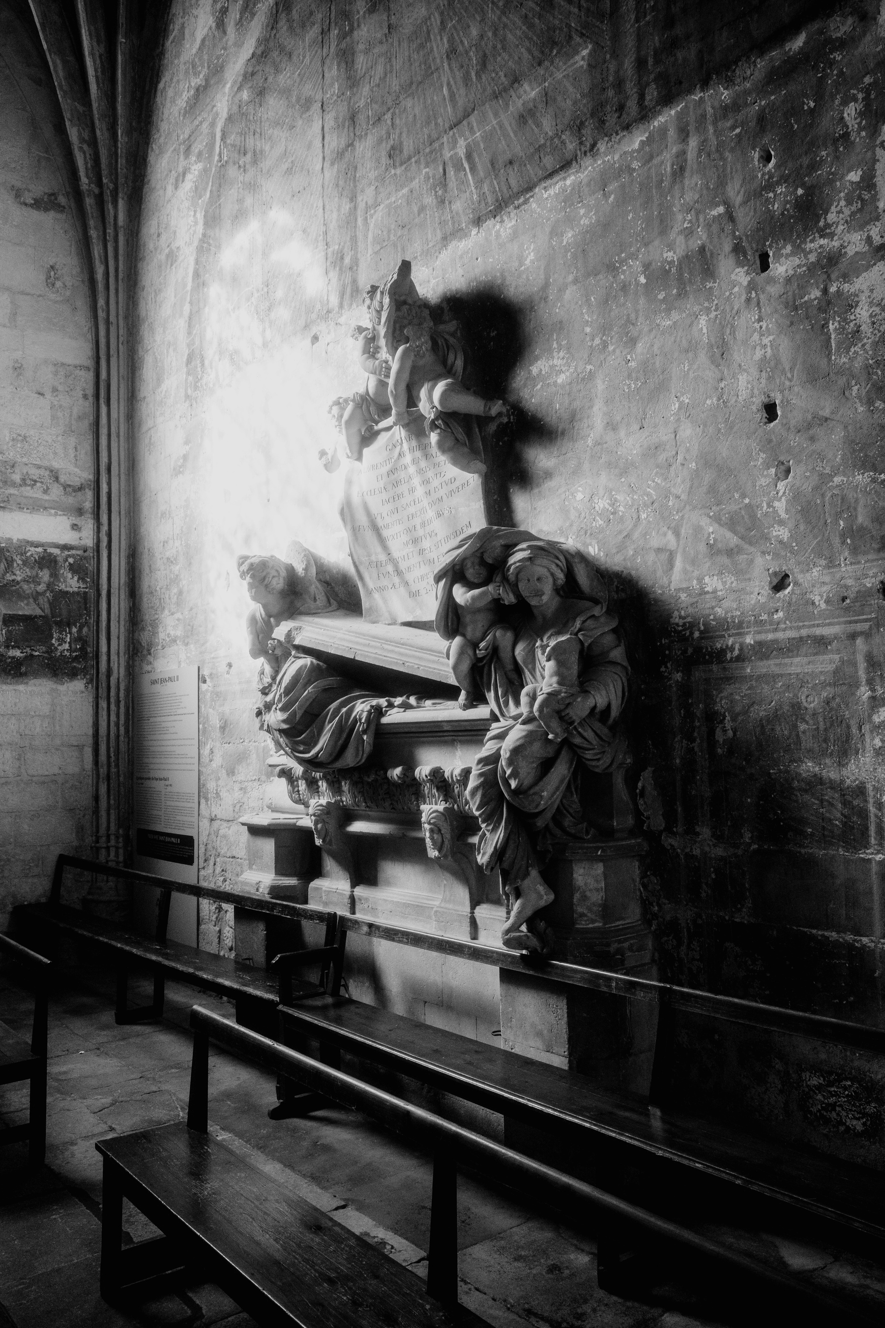 The image shows a black-and-white photograph of a detailed and dramatic stone tomb adorned with sculptures of figures, located against a textured stone wall in a dimly lit church interior.