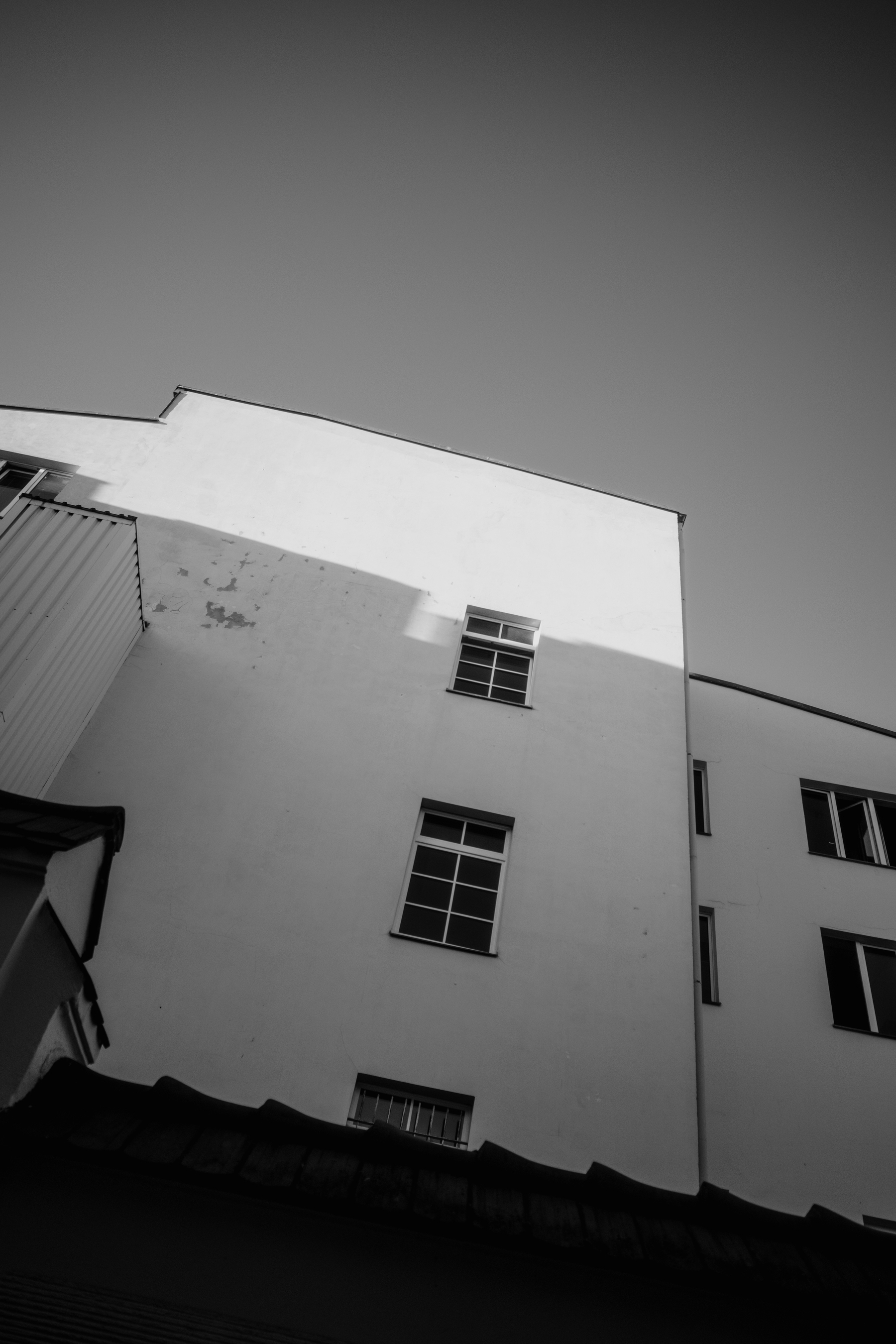 The image is a black-and-white photo of a building's exterior wall, viewed from below, with a few windows and sharp shadows cast by the sun.