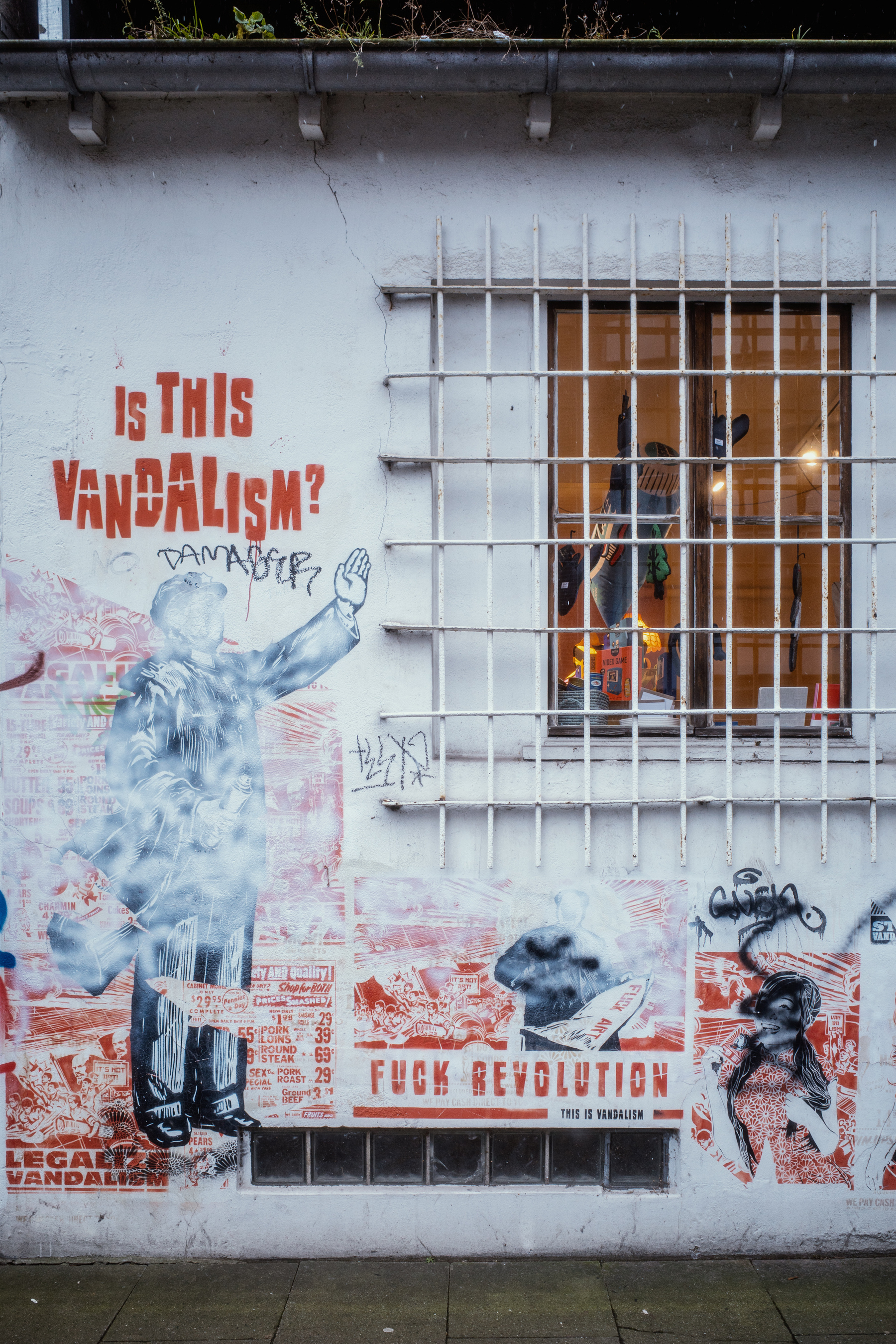 The image depicts a wall covered with graffiti and posters featuring provocative text such as “Is this vandalism?” and “FUCK REVOLUTION,” alongside a barred window revealing a lit interior with various objects.