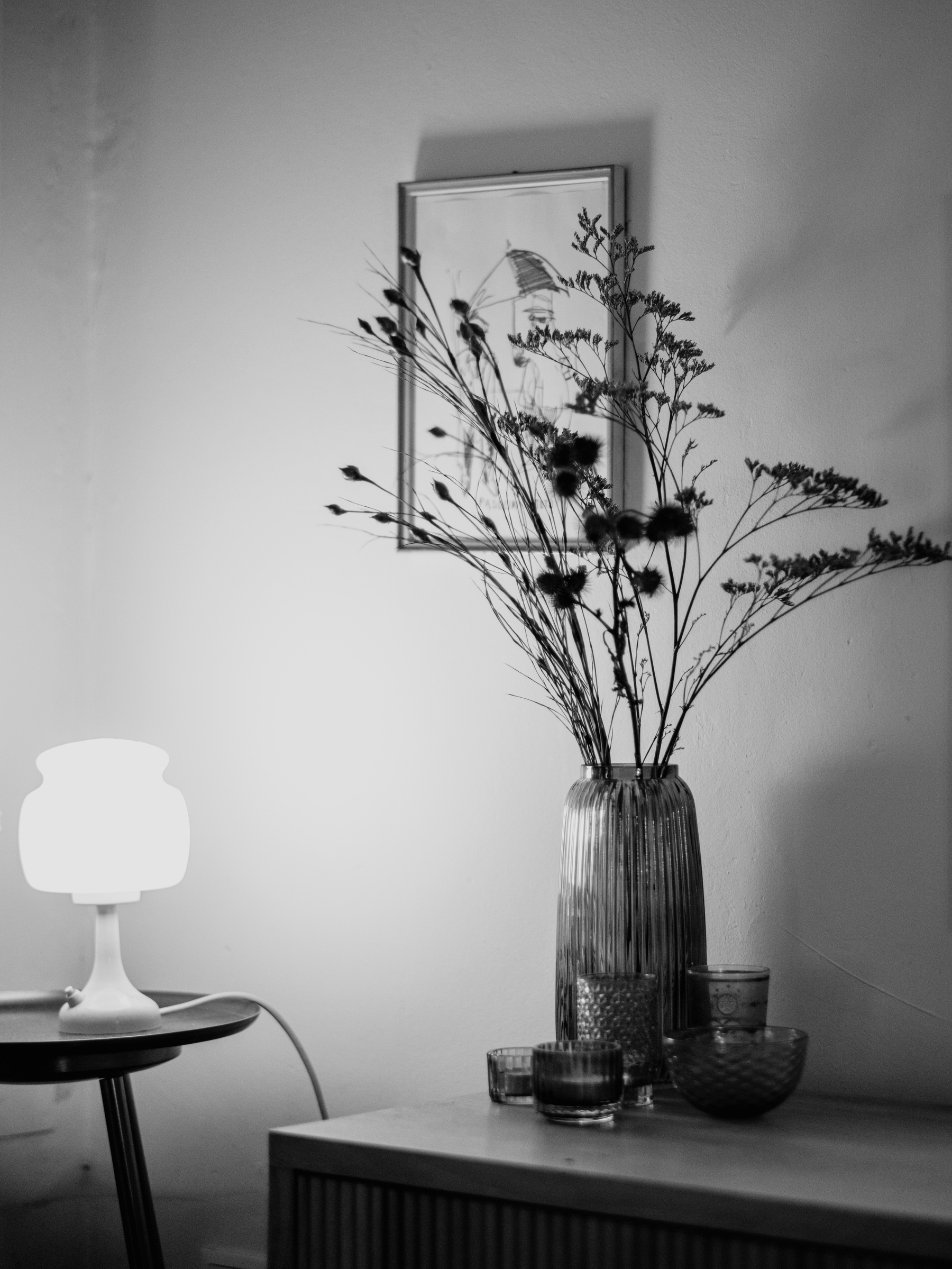 The black-and-white image shows a minimalist interior scene with a lit table lamp on a small table, a vase with tall dried flowers, and several decorative glass containers on a wooden surface, framed by a simple artwork on the wall.