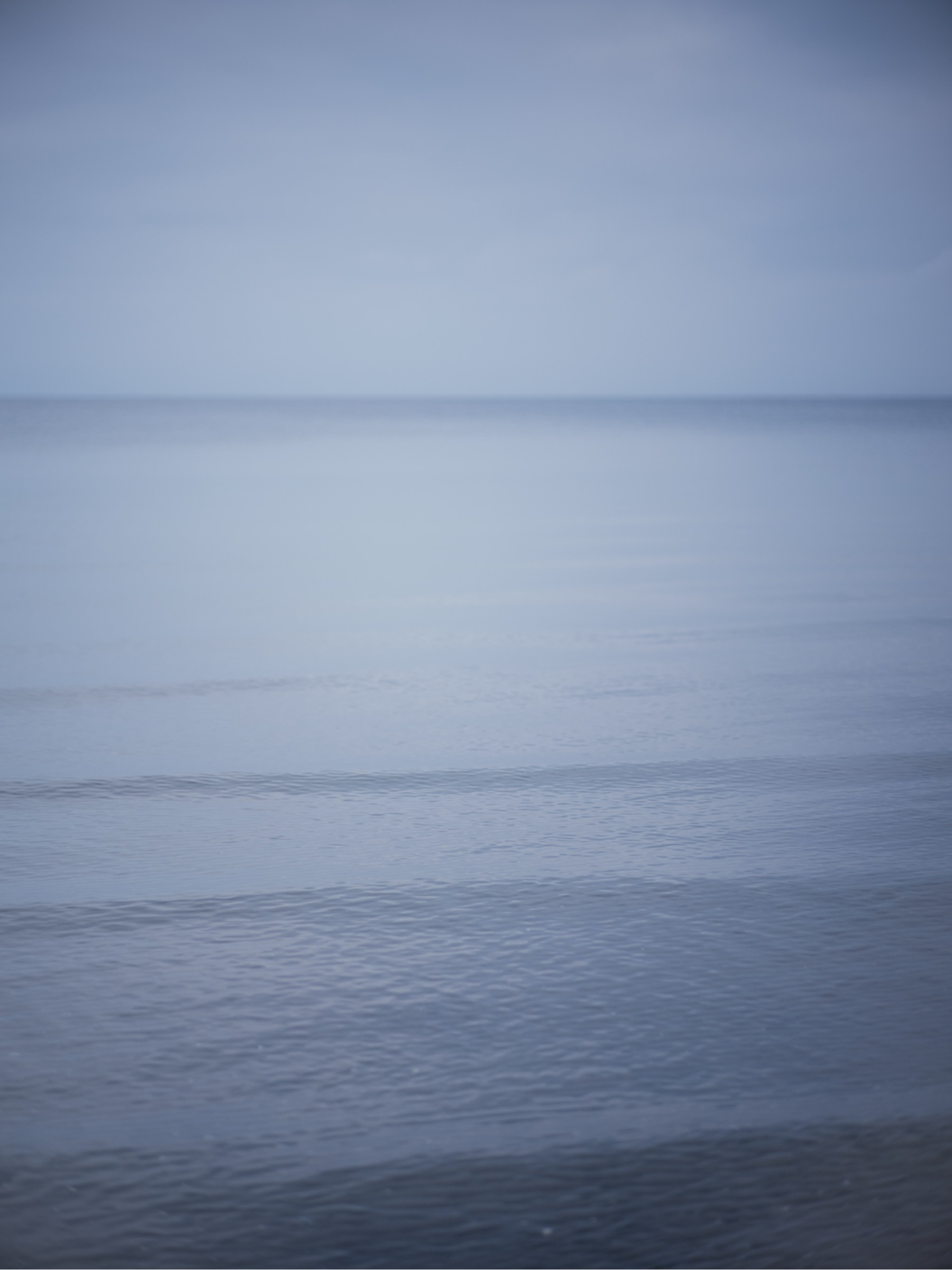 A calm, softly lit seascape shows gentle ripples on a blue-gray ocean beneath an overcast sky.