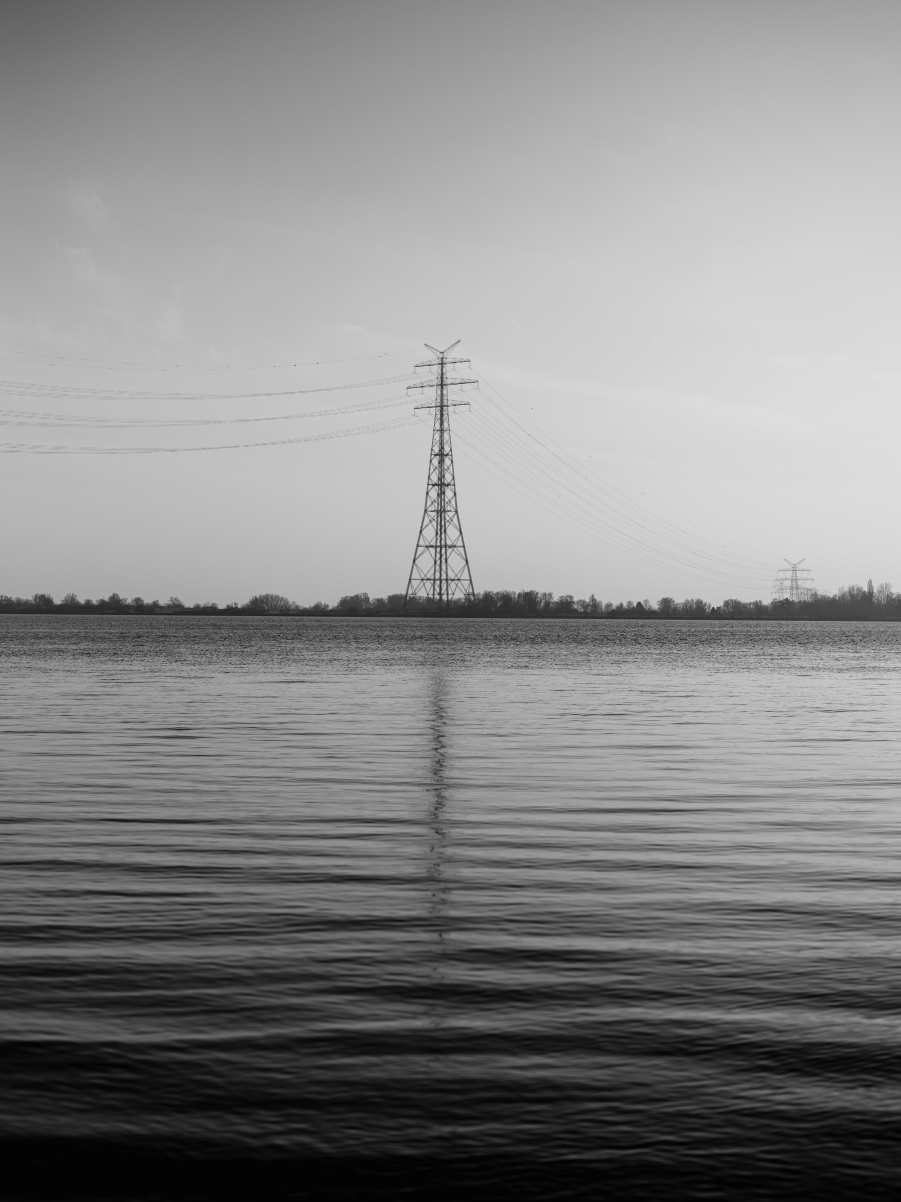 A tall electricity pylon stands near a calm body of water, its reflection rippling on the surface, with power lines extending into the distance under a vast sky.