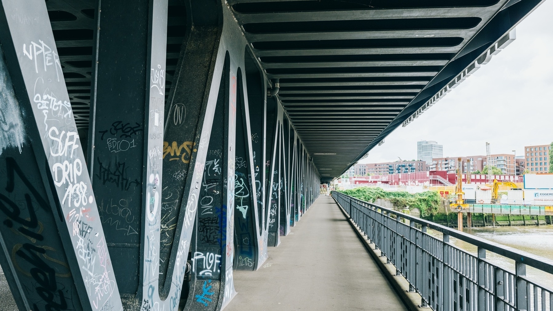 The image shows a long pedestrian walkway under a steel bridge, with graffiti-covered beams on the left and a riverside construction site with modern buildings on the right.