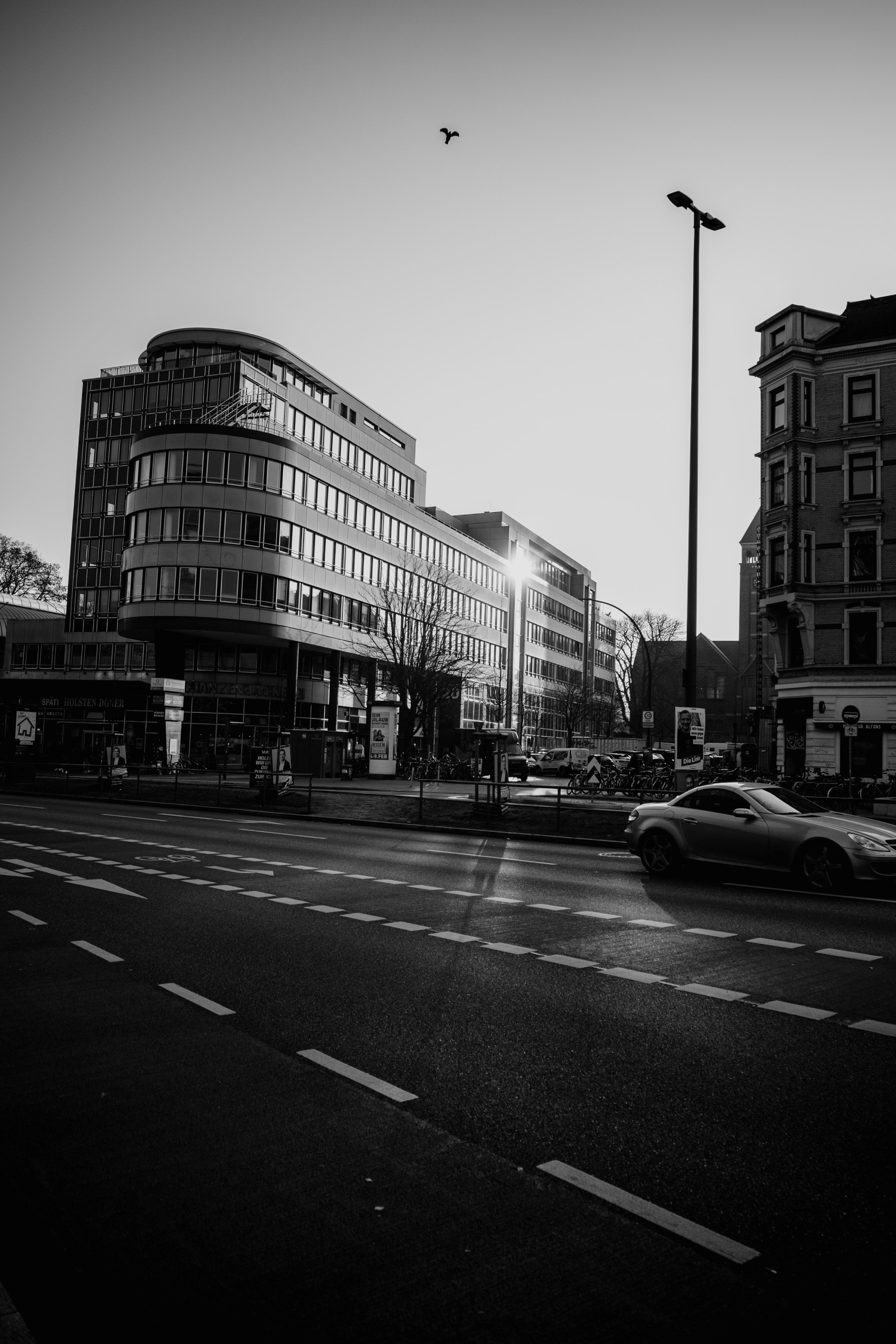 This black and white photograph captures an urban street scene with a modern, curved glass-fronted building, a classic brick structure, a moving car, a bird in flight, and the sun reflecting off one of the windows, creating a striking highlight.