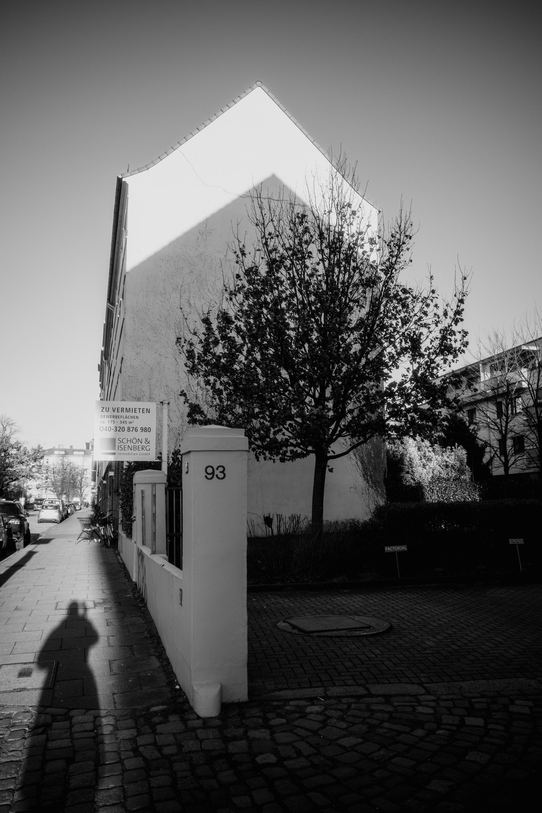 The black and white photograph captures a quiet urban street scene featuring a starkly lit building with a dramatic triangular shadow, a tree partially obscuring the facade, and a person’s shadow stretching onto the sidewalk in the foreground.