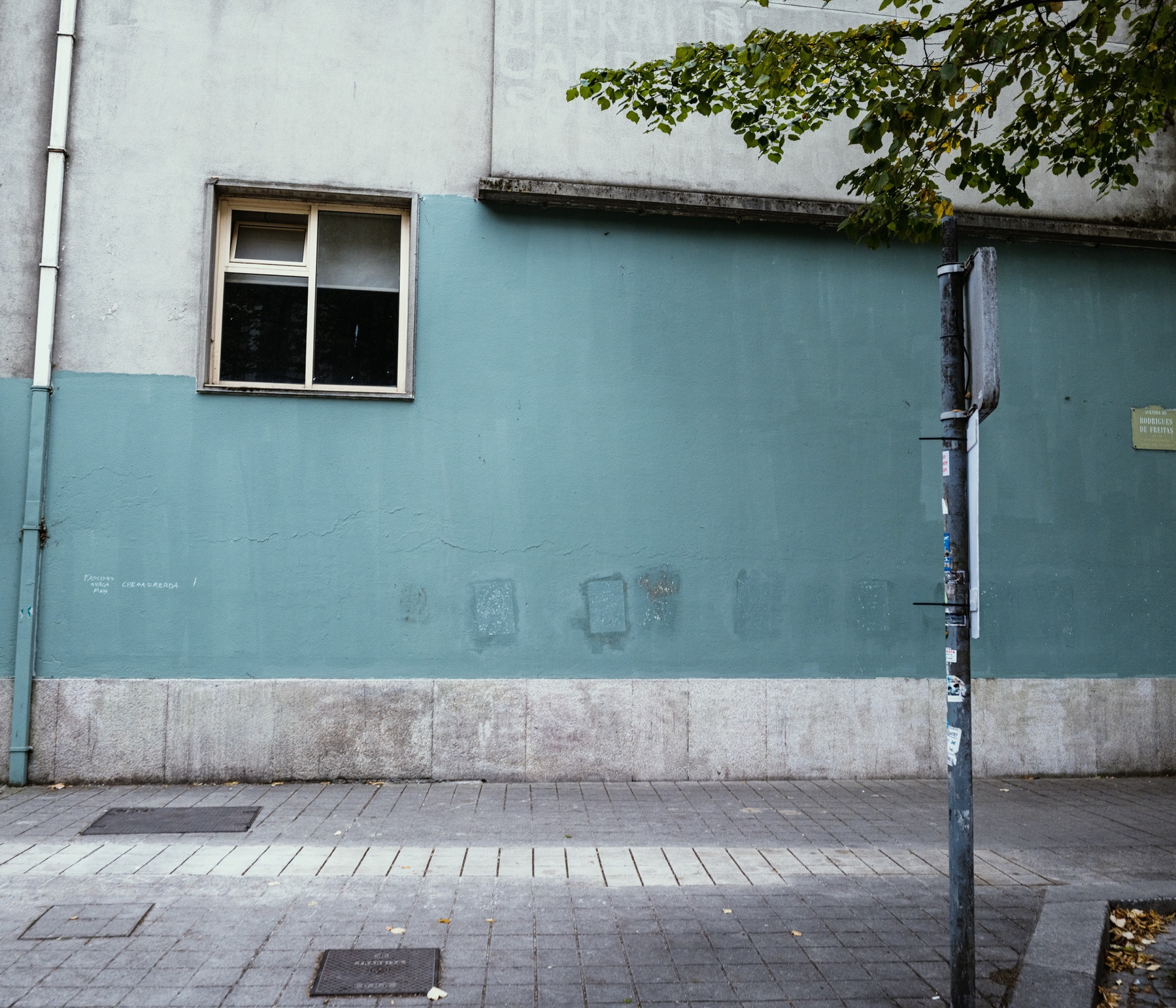 A quiet urban street scene shows a pale blue and gray building wall with a single window, a weathered signpost, and a few tree branches extending overhead.