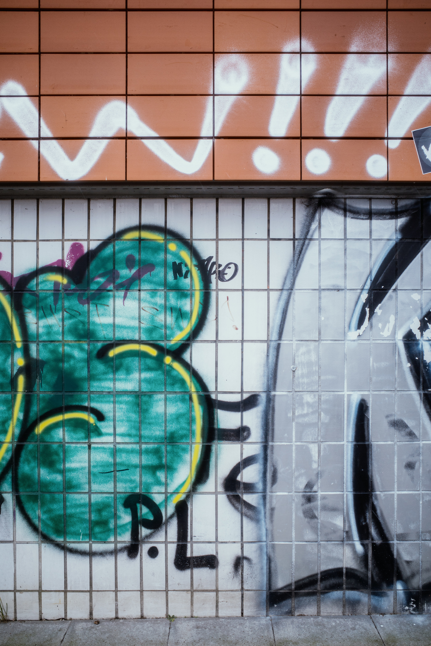 The image shows a tiled wall with colorful graffiti, including a green shape outlined in yellow and white spray-painted markings above.