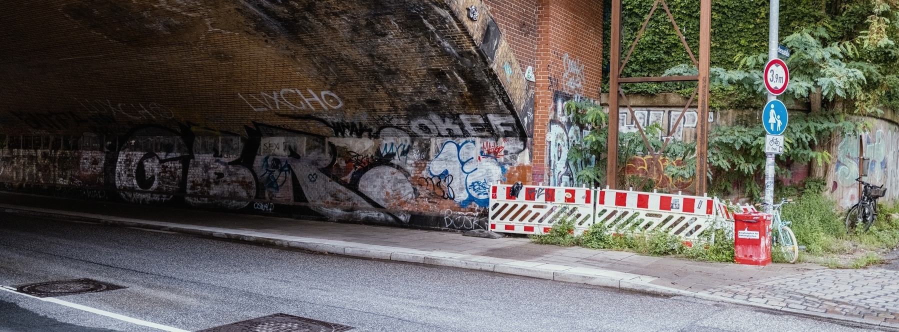 The image shows a graffiti-covered brick bridge underpass beside a street, with construction barriers, traffic signs, and overgrown vegetation in the background.