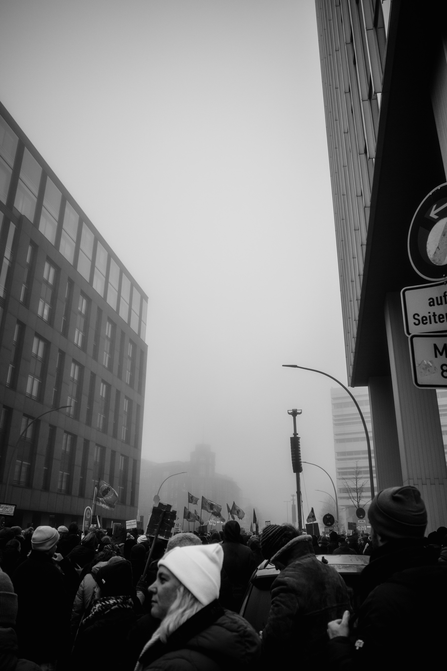 The black and white photograph depicts a foggy urban protest scene with a dense crowd carrying flags and signs, framed by towering modern buildings and street signs, creating a sense of movement and tension.