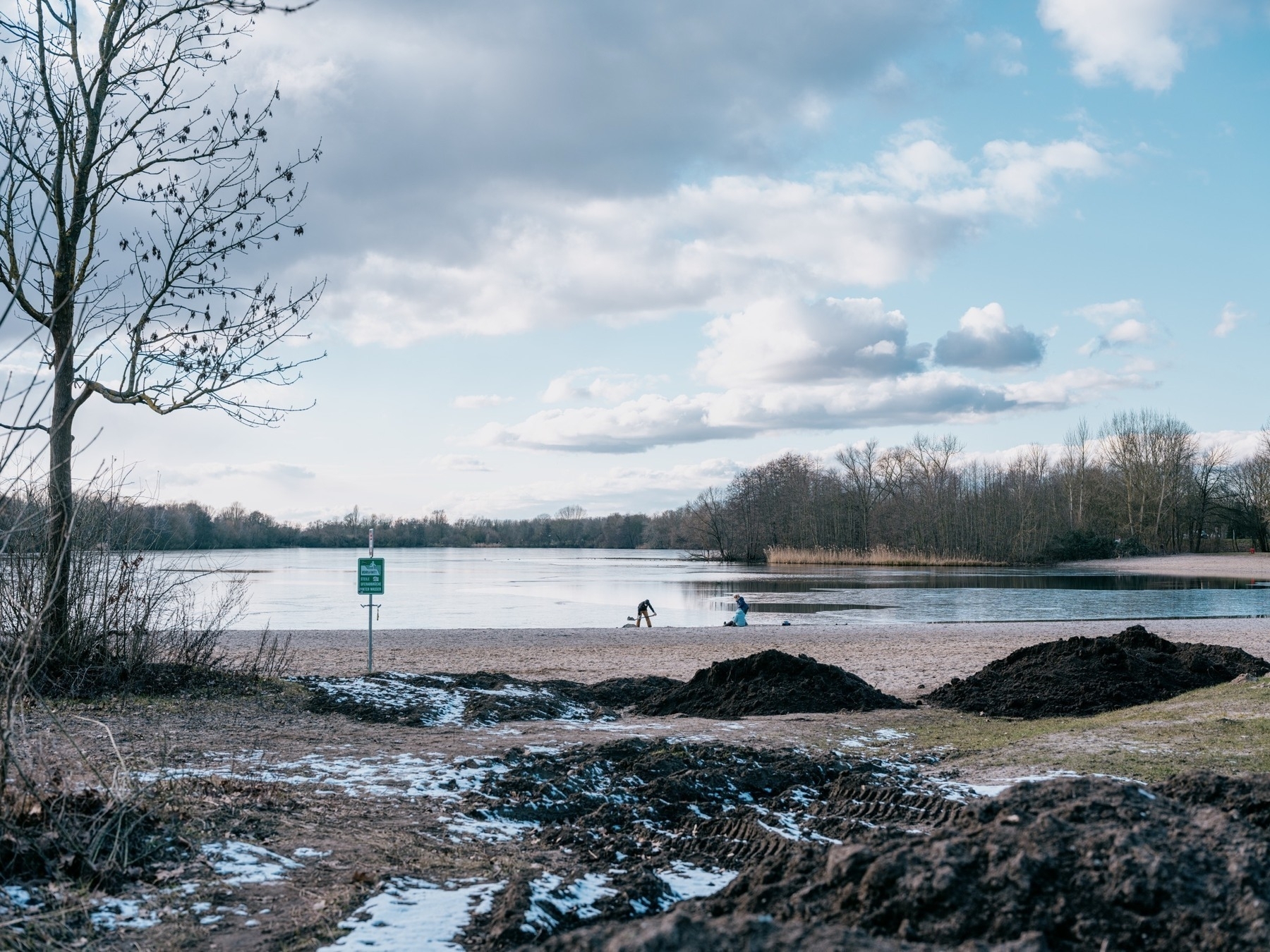 A serene lake view in late winter or early spring, with bare trees lining the shore, patches of melting snow on muddy ground, and people in the distance on a small beach by the water under a cloudy blue sky.​​​​​​​​​​​​​​​​