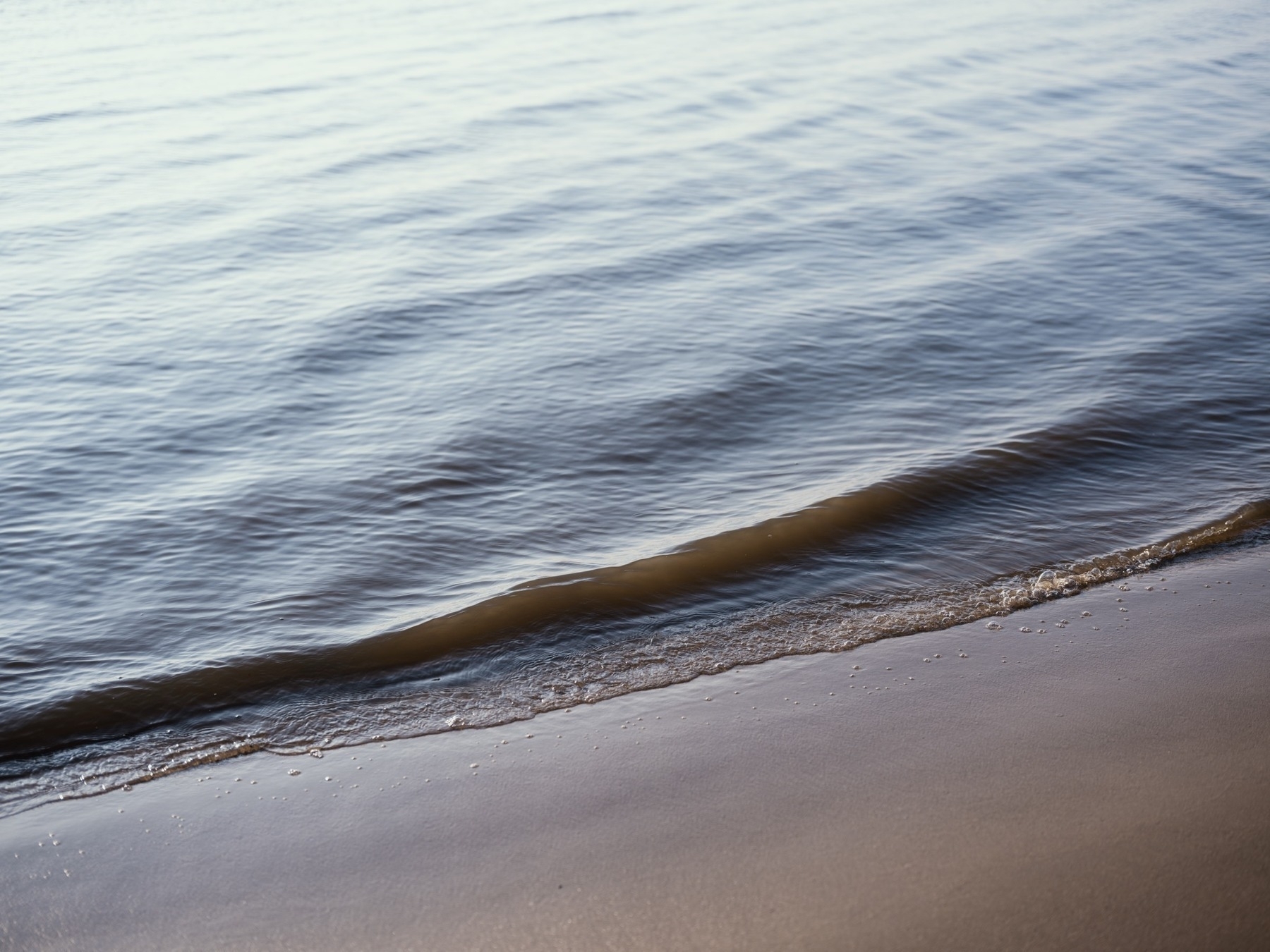 This photograph captures a gentle wave rolling onto a smooth sandy shore, with soft ripples in the water reflecting natural light, creating a calm and peaceful scene.