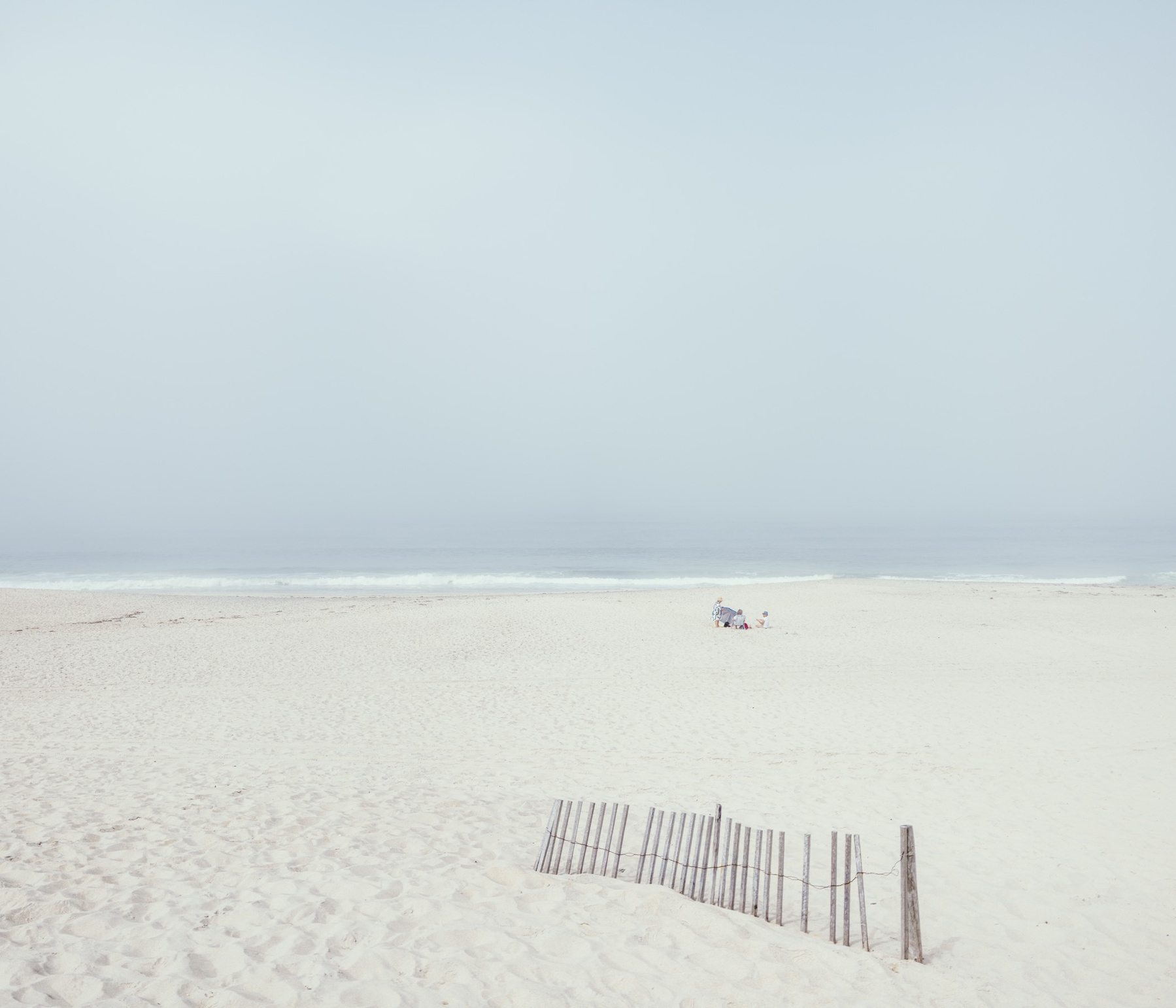 A misty, almost empty beach stretches toward the calm sea, with a small group of people sitting near the shoreline behind a weathered sand fence.