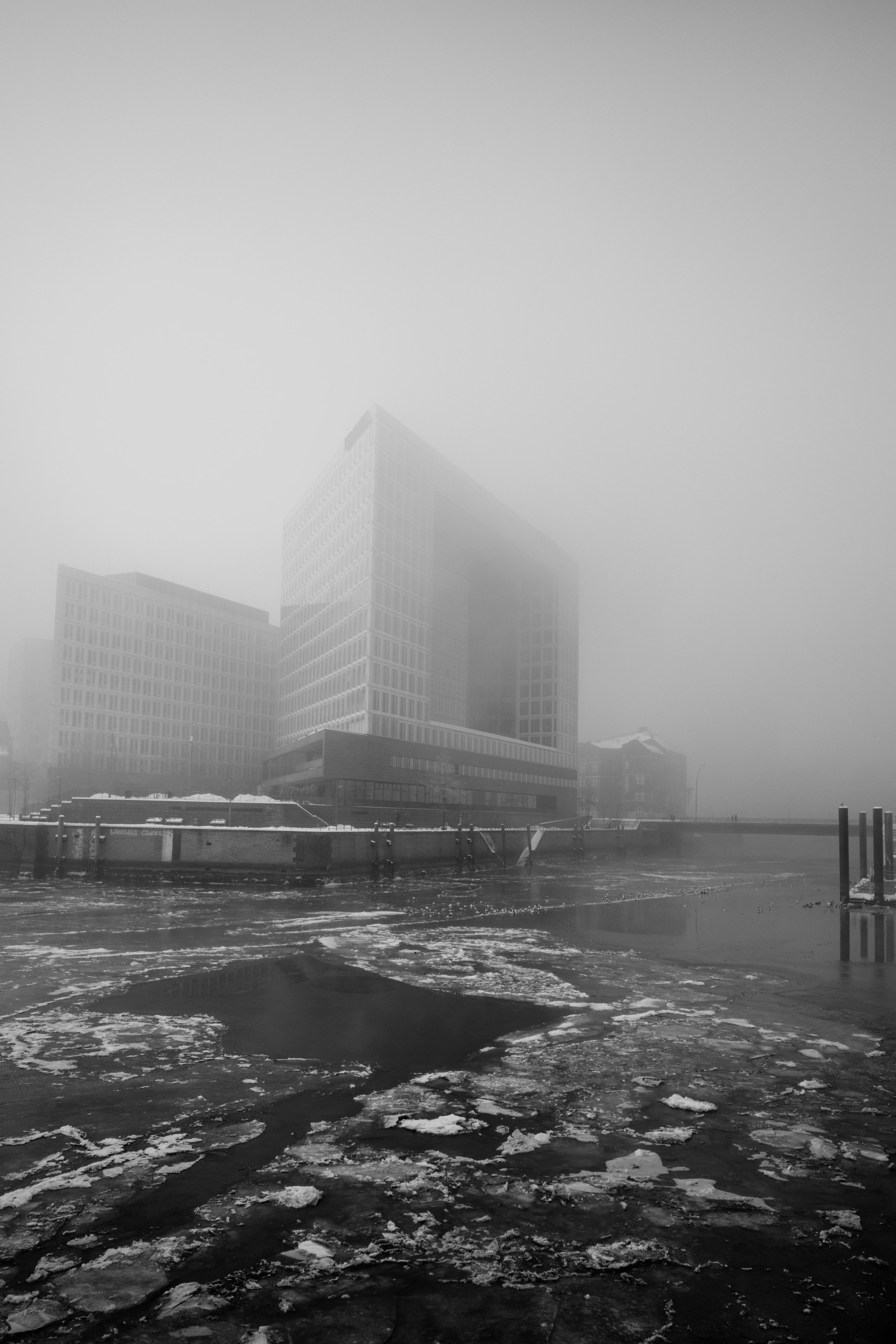A fog-shrouded modern building complex rises beside a partially frozen body of water, creating a stark, minimalist urban scene.