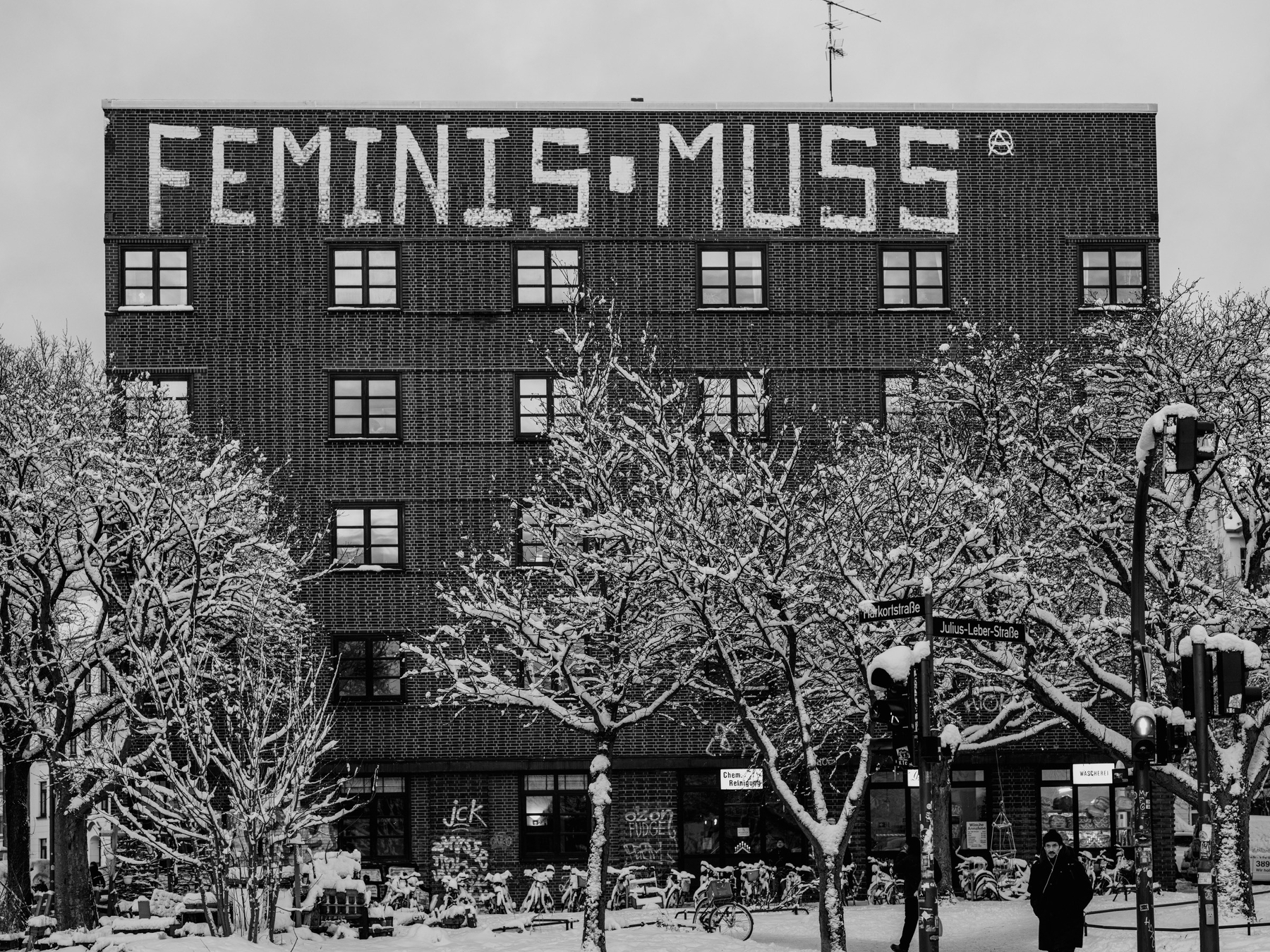 A black-and-white photograph shows a brick apartment building with the large rooftop graffiti “FEMINIS-MUSS,” framed by snow-covered trees and a wintery street intersection below.