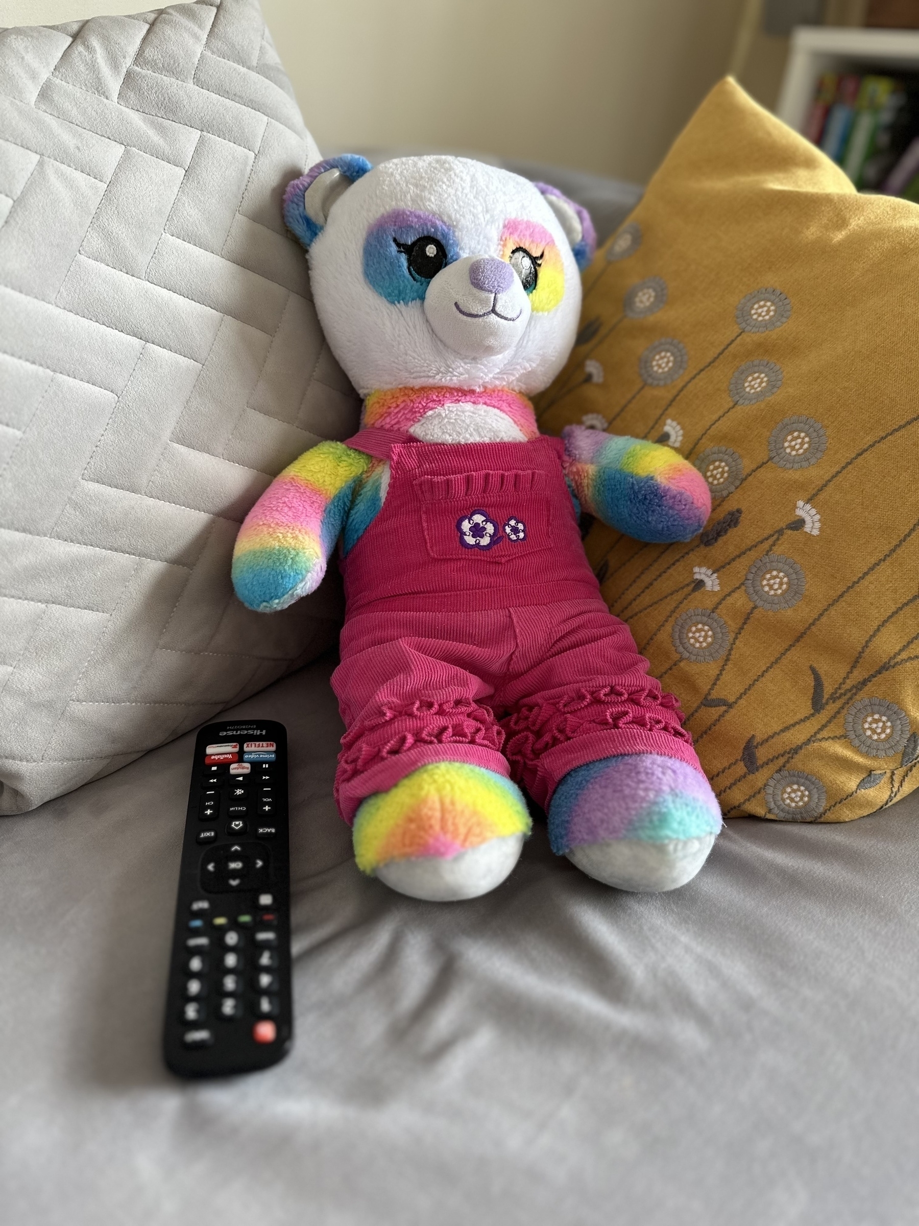 Teddy bear sat on the sofa, holding a TV remote.
