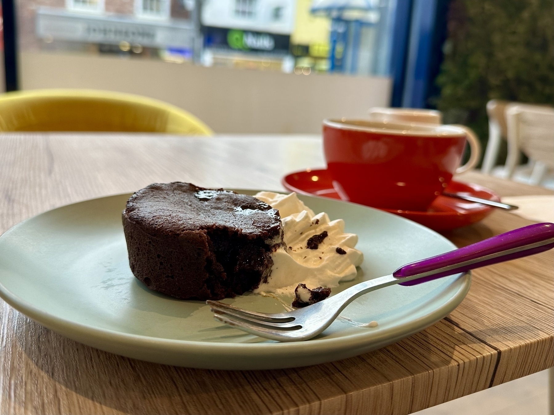 A chocolate dessert with whipped cream and a fork is placed on a green plate, accompanied by a red cup and saucer on a wooden table.