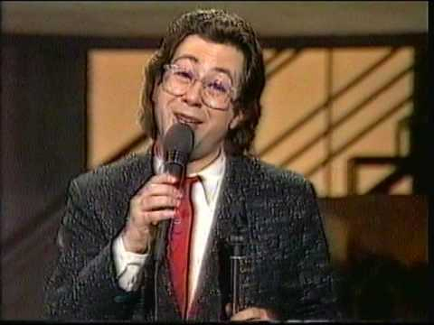 Ben Elton, back in the 1980s, holding a microphone and talking to camera.