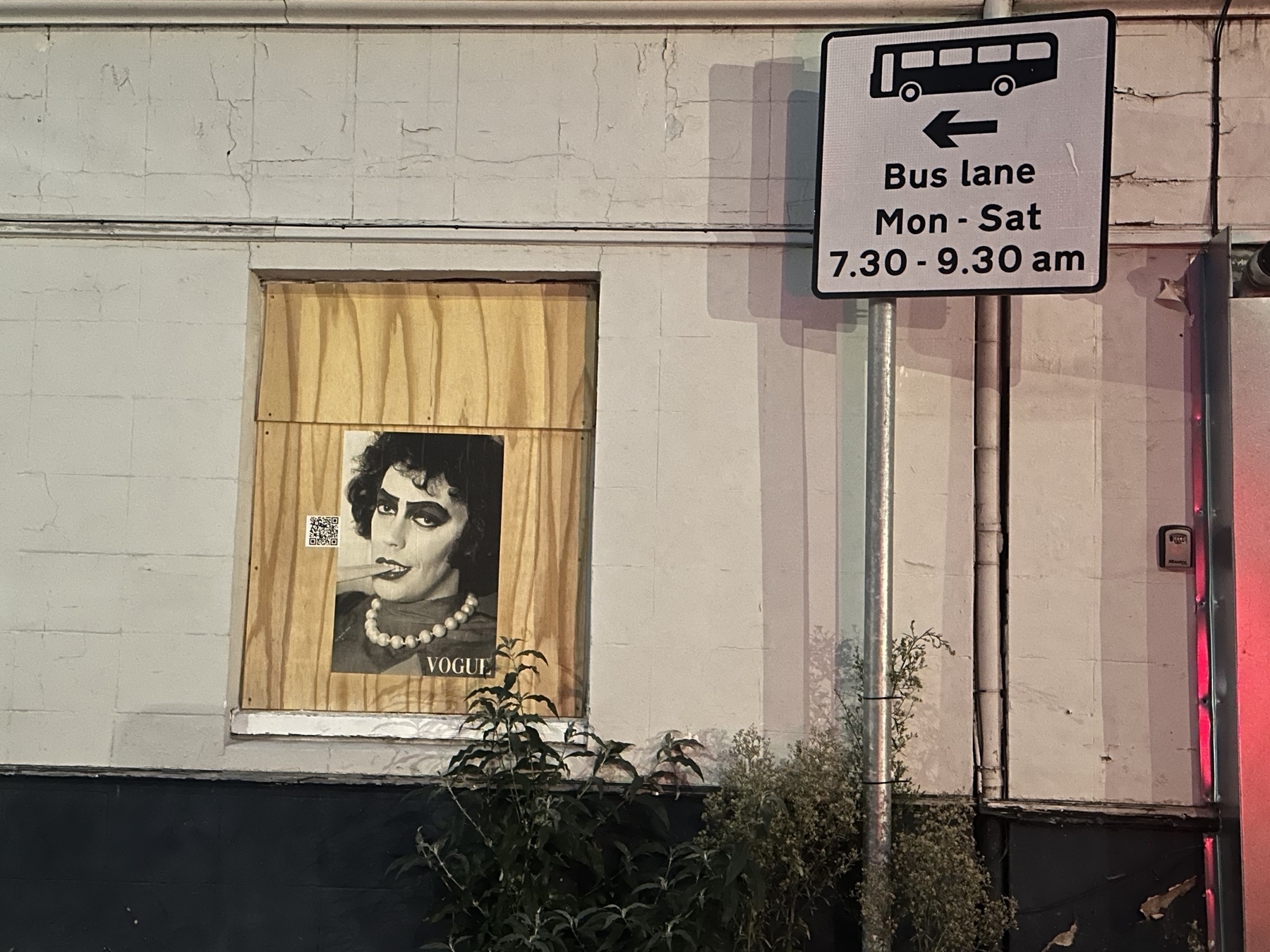 A street sign indicating bus lane times is next to a wall with a poster of a person displaying a dramatic makeup look and the word VOGUE below.