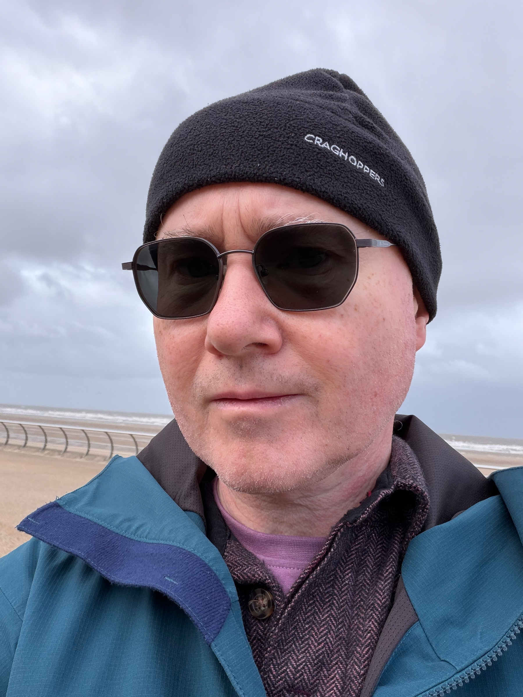 Me wearing dark prescription glasses, a black beanie, and a blue jacket and standing outdoors near a beach with cloudy skies overhead.