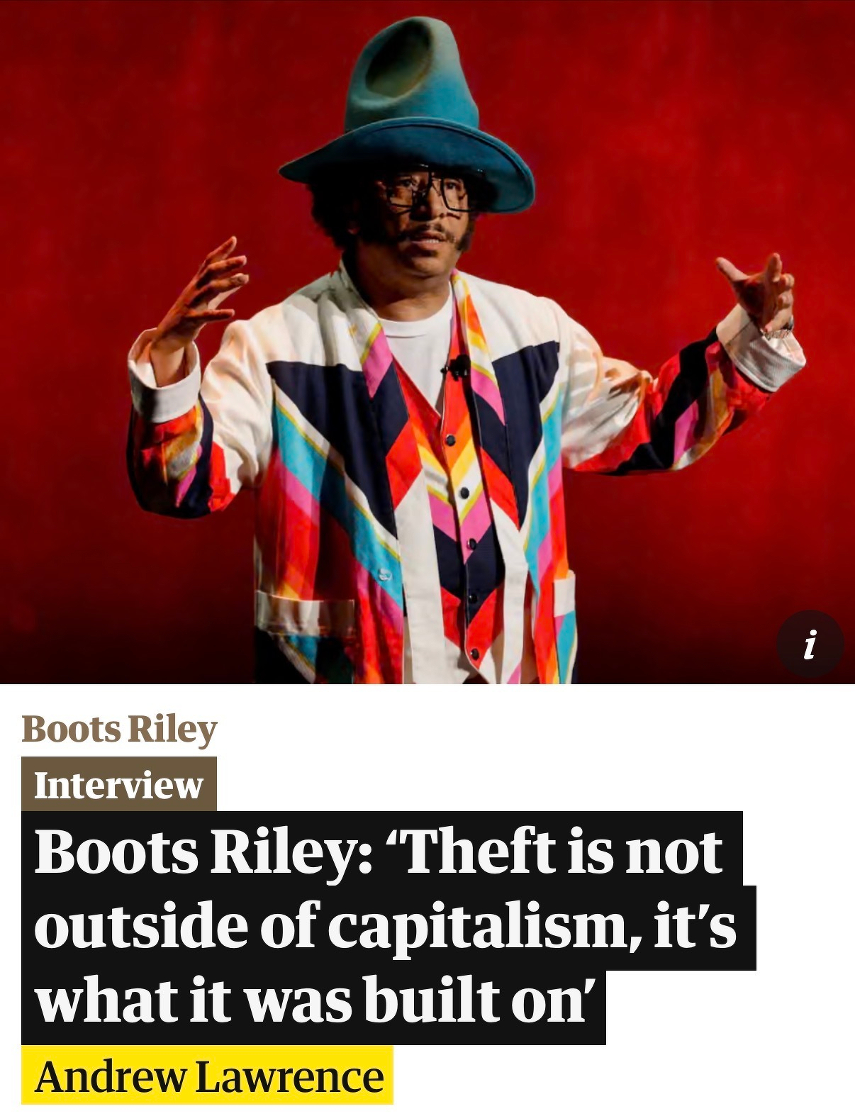 Boots Riley wearing a colorful jacket and hat is standing in front of a red background.