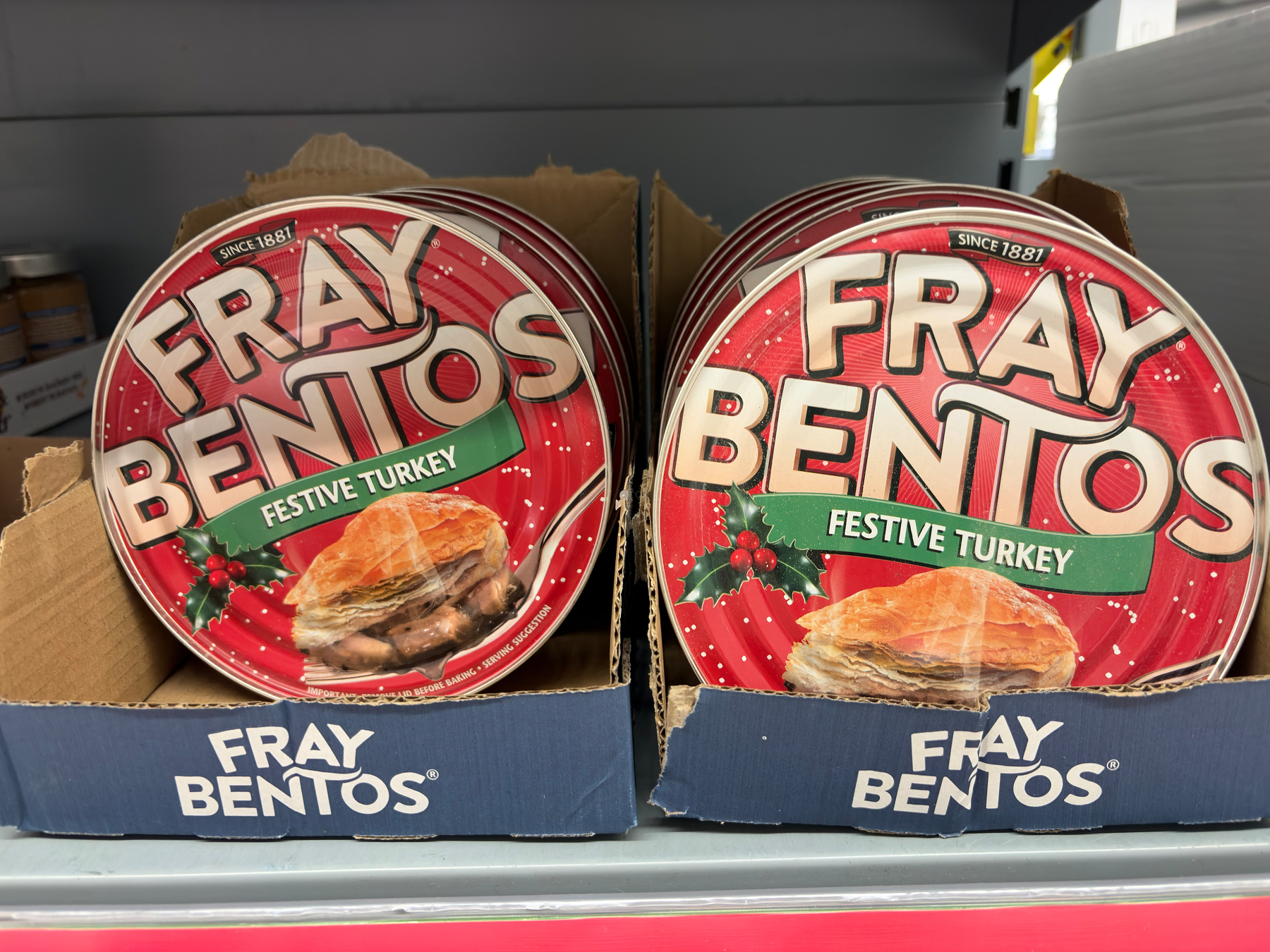 Two tins of Fray Bentos Festive Turkey meat pies are displayed in a cardboard box on a store shelf.