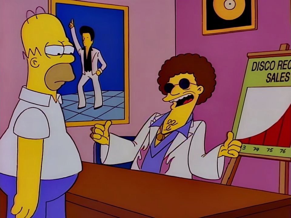 A screen grab from The Simpsons showing Disco Stu with his thumbs up and smiling. M