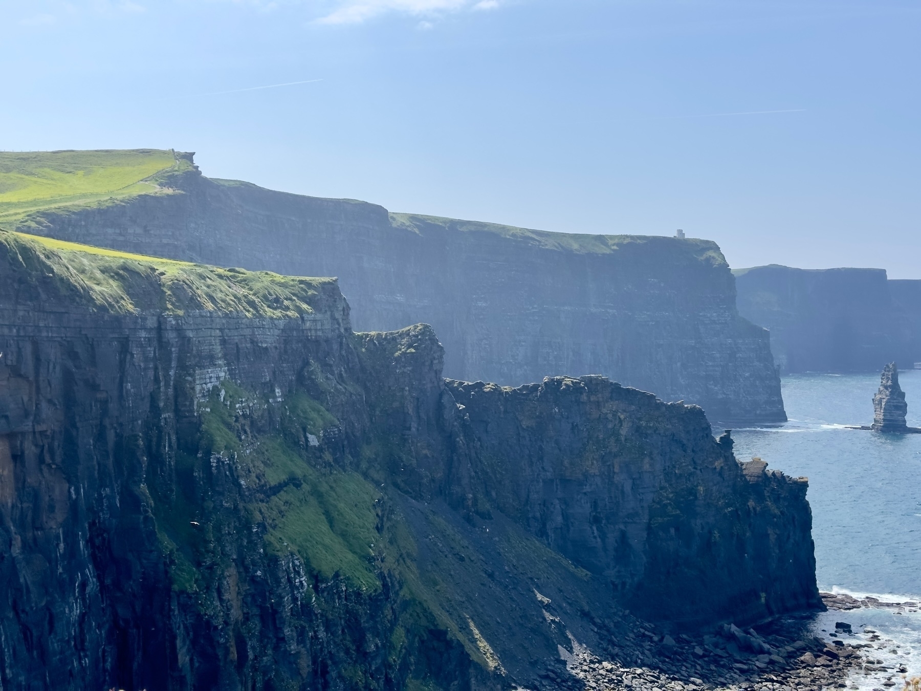 Auto-generated description: Majestic sea cliffs, covered in lush green grass, rise dramatically above the ocean, with rocky outcrops and a faint blue sky in the backdrop.
