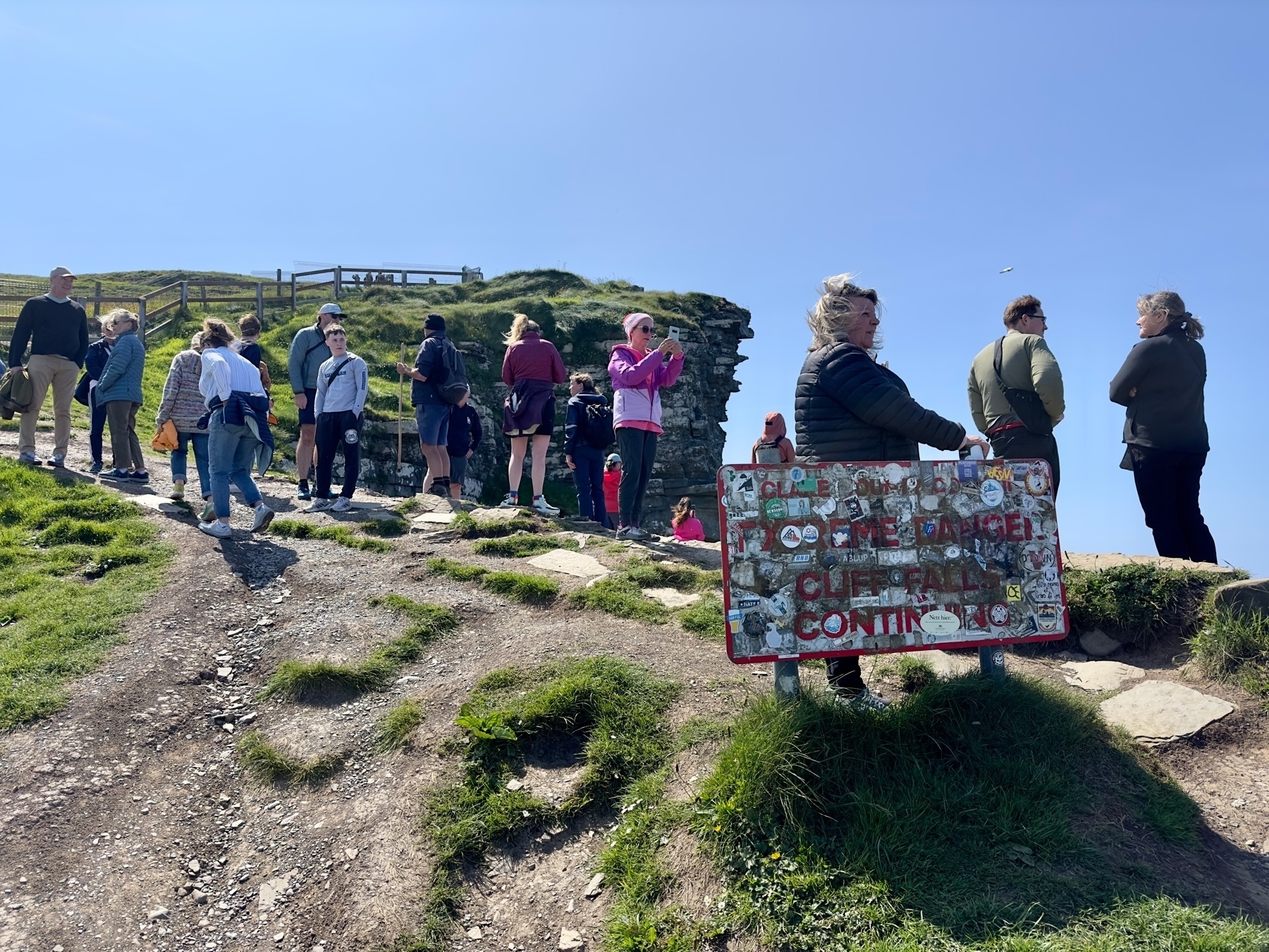 Auto-generated description: A group of people are gathered outdoors on a rocky path near a cliff, with one person holding a sign warning about dangers.