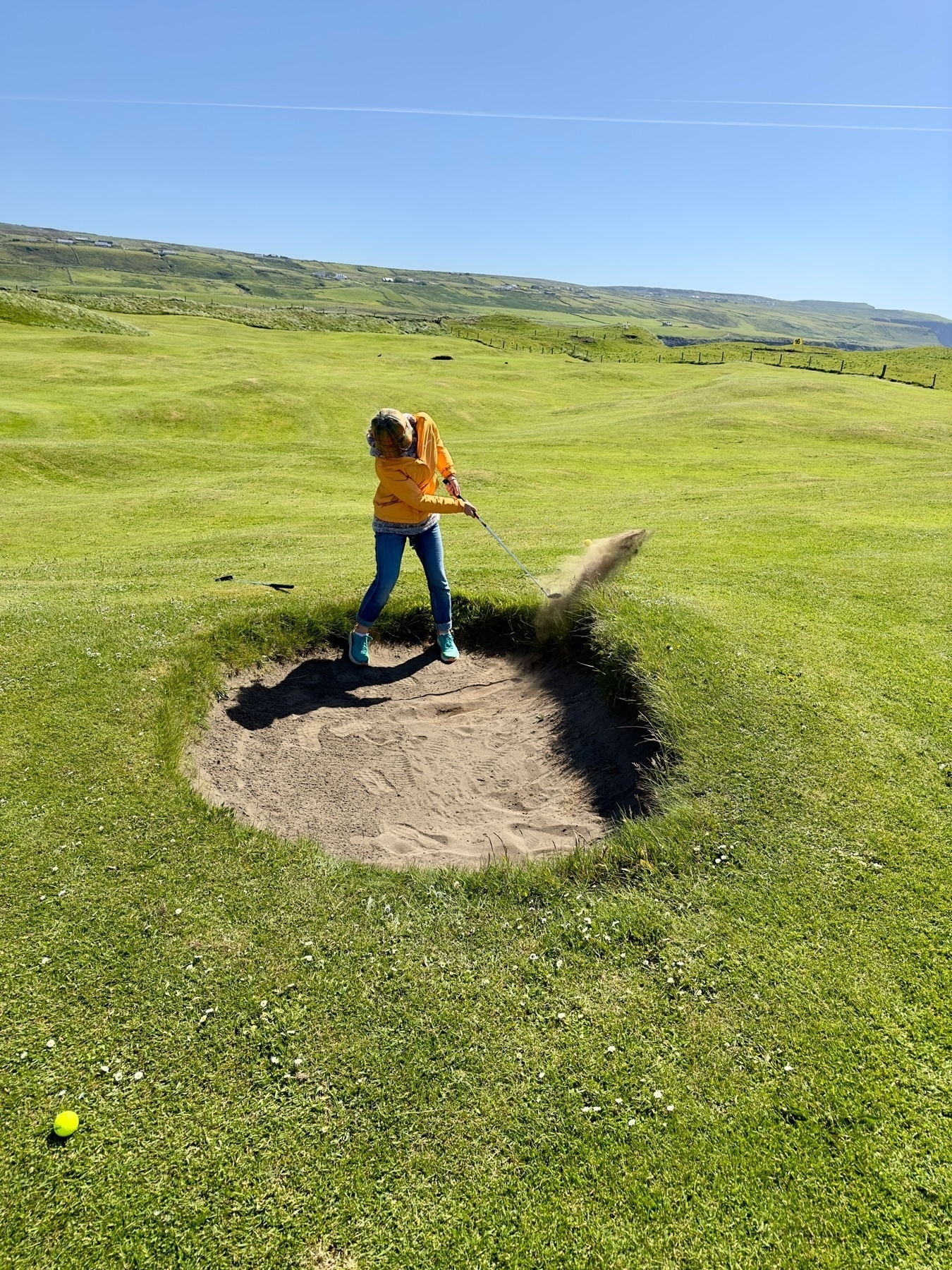 Auto-generated description: A person in a yellow shirt and blue jeans is hitting a golf ball out of a sand trap on a grassy golf course.