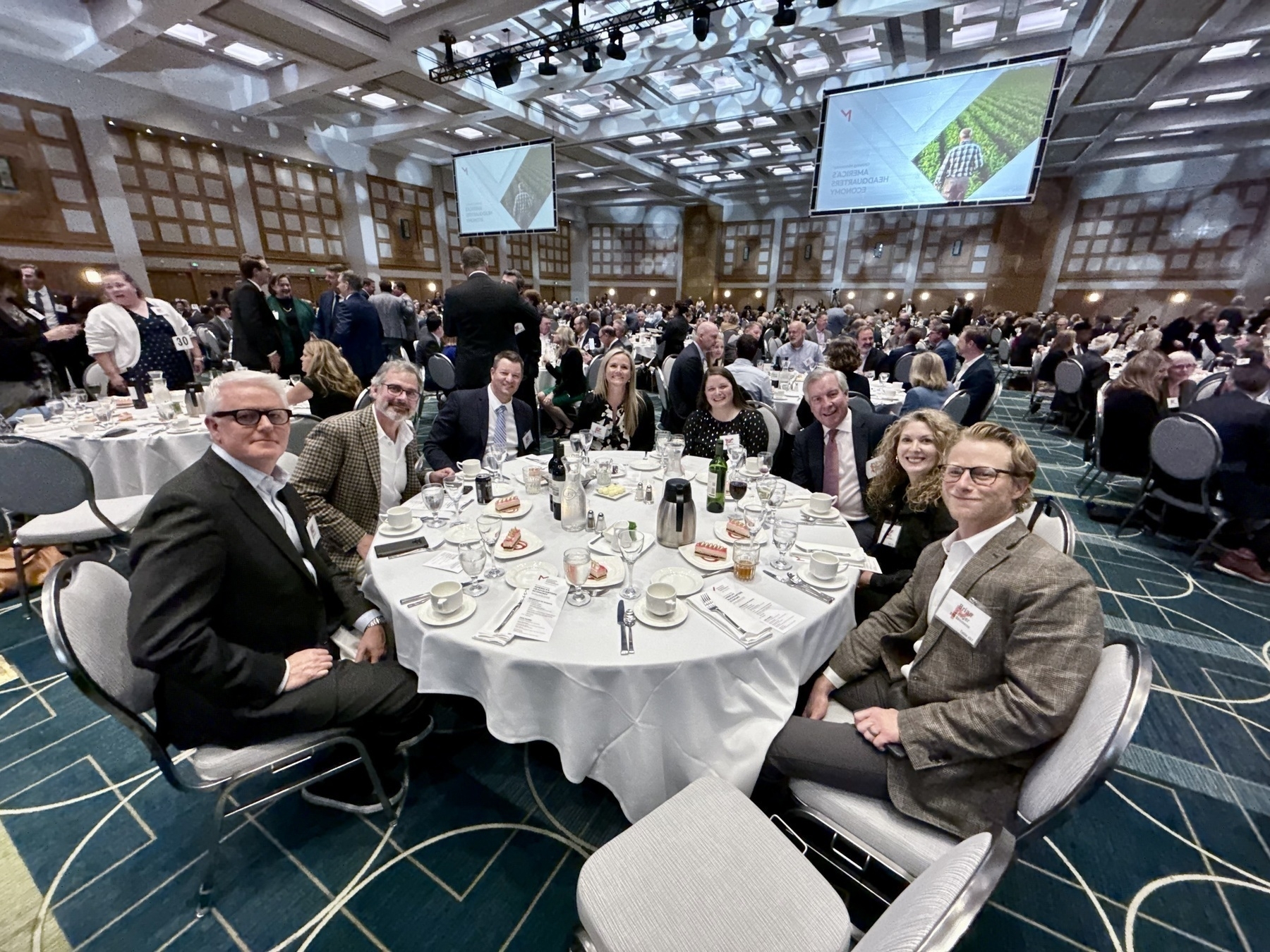 Auto-generated description: A group of people are seated around a table in a large, crowded banquet hall with a formal setting.