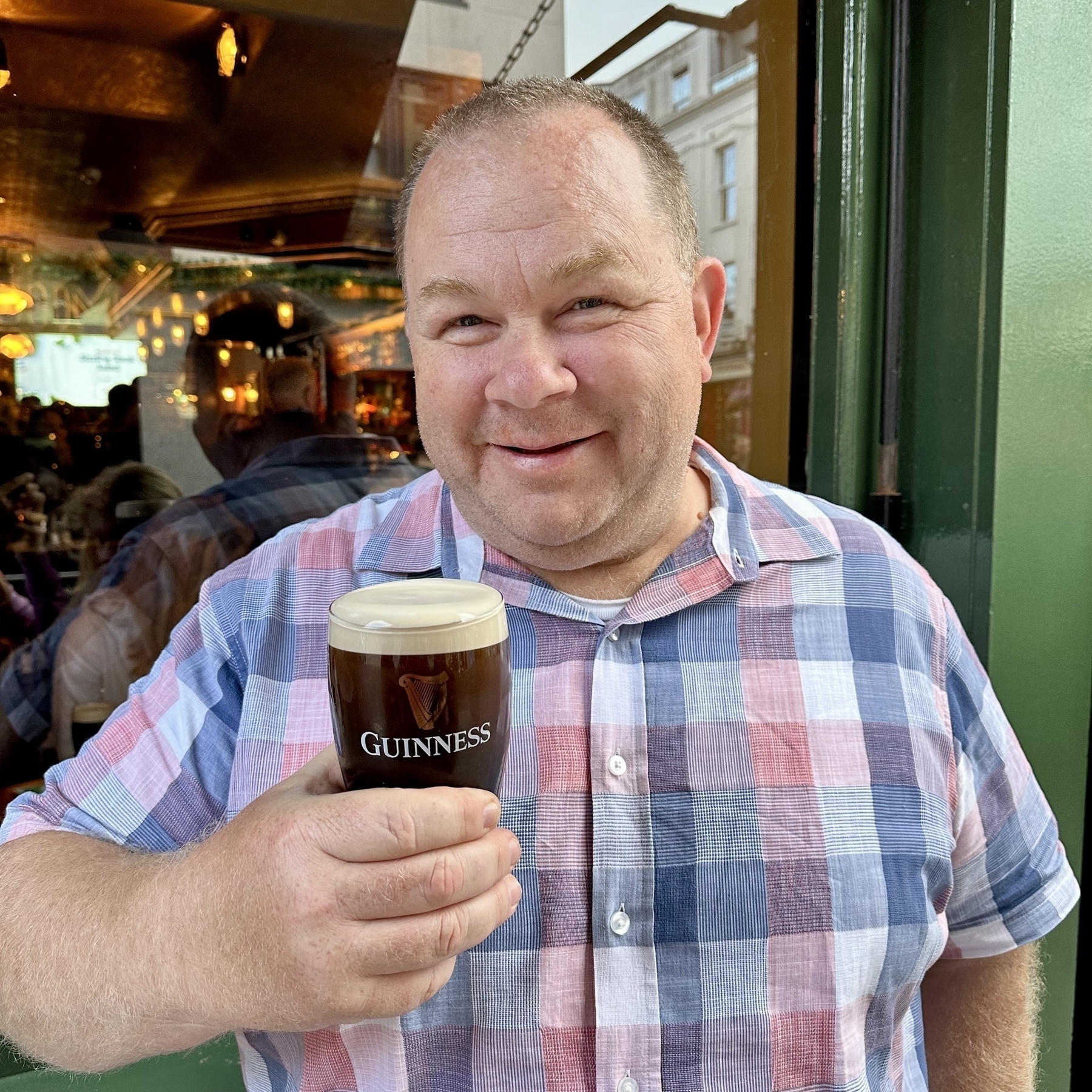 Auto-generated description: A smiling man holds a pint of Guinness while standing in front of a pub.
