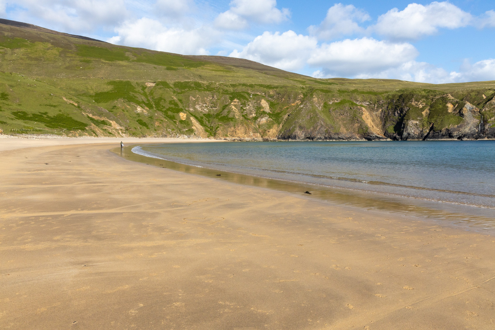 Auto-generated description: A deserted sandy beach with clear blue water is backed by green, rocky hills under a partly cloudy sky.