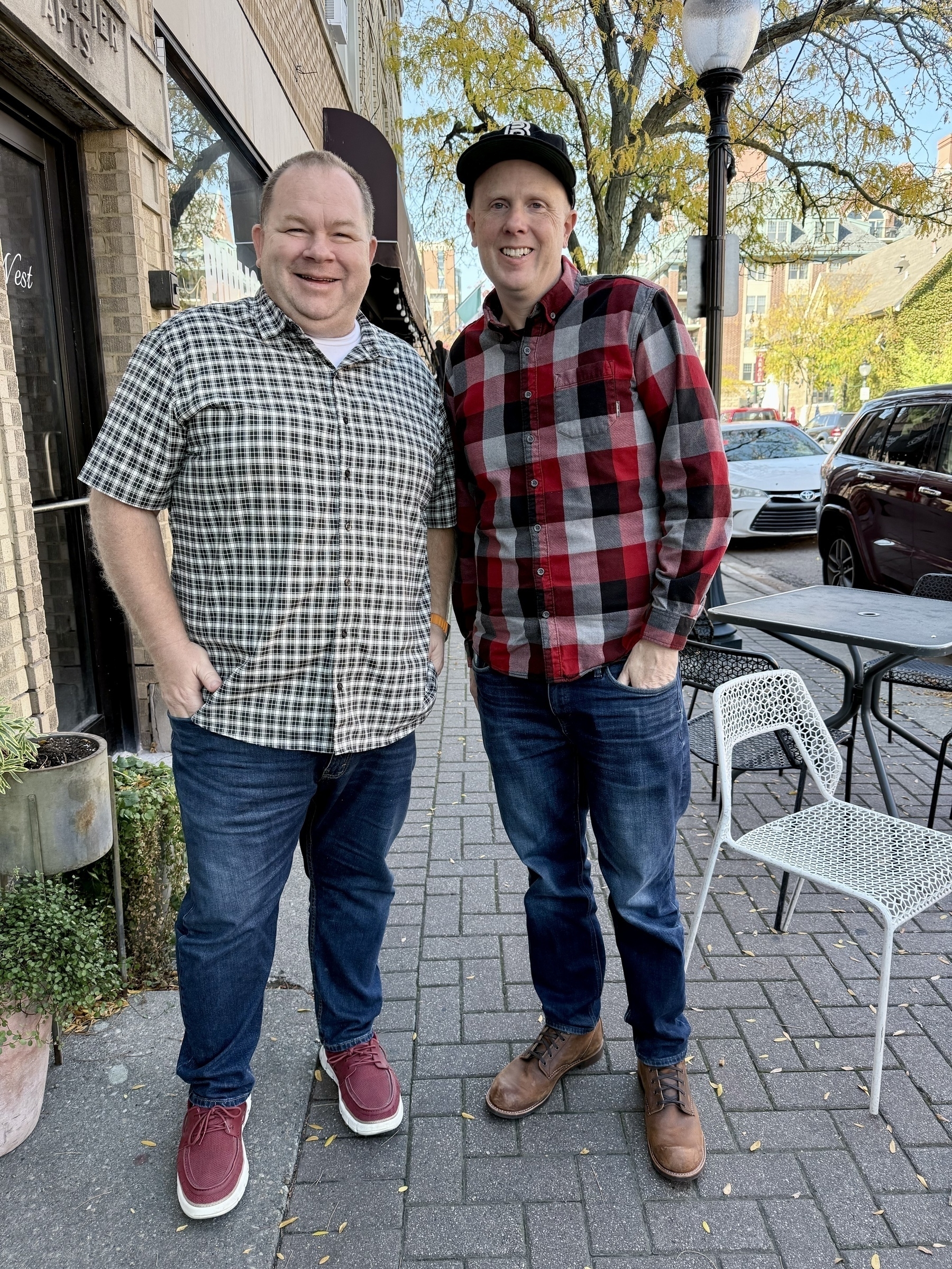 Auto-generated description: Two people are standing on a sidewalk outside, smiling at the camera, with trees and a few parked cars in the background.