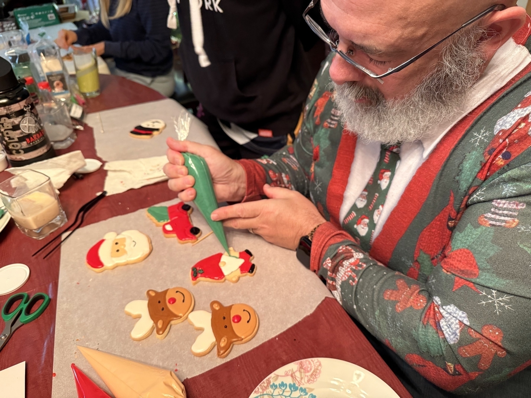 Auto-generated description: A person decorates holiday-themed cookies with icing at a table, featuring designs like Santa and reindeer.