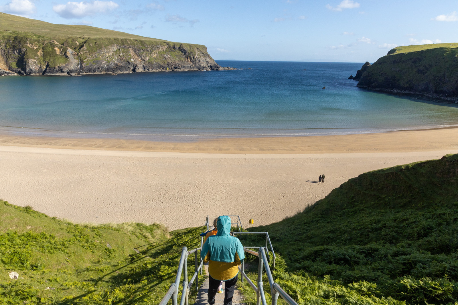 Auto-generated description: A person wearing a colorful jacket walks down a staircase towards a pristine, sandy beach bordered by green hills and overlooking a calm, blue bay.