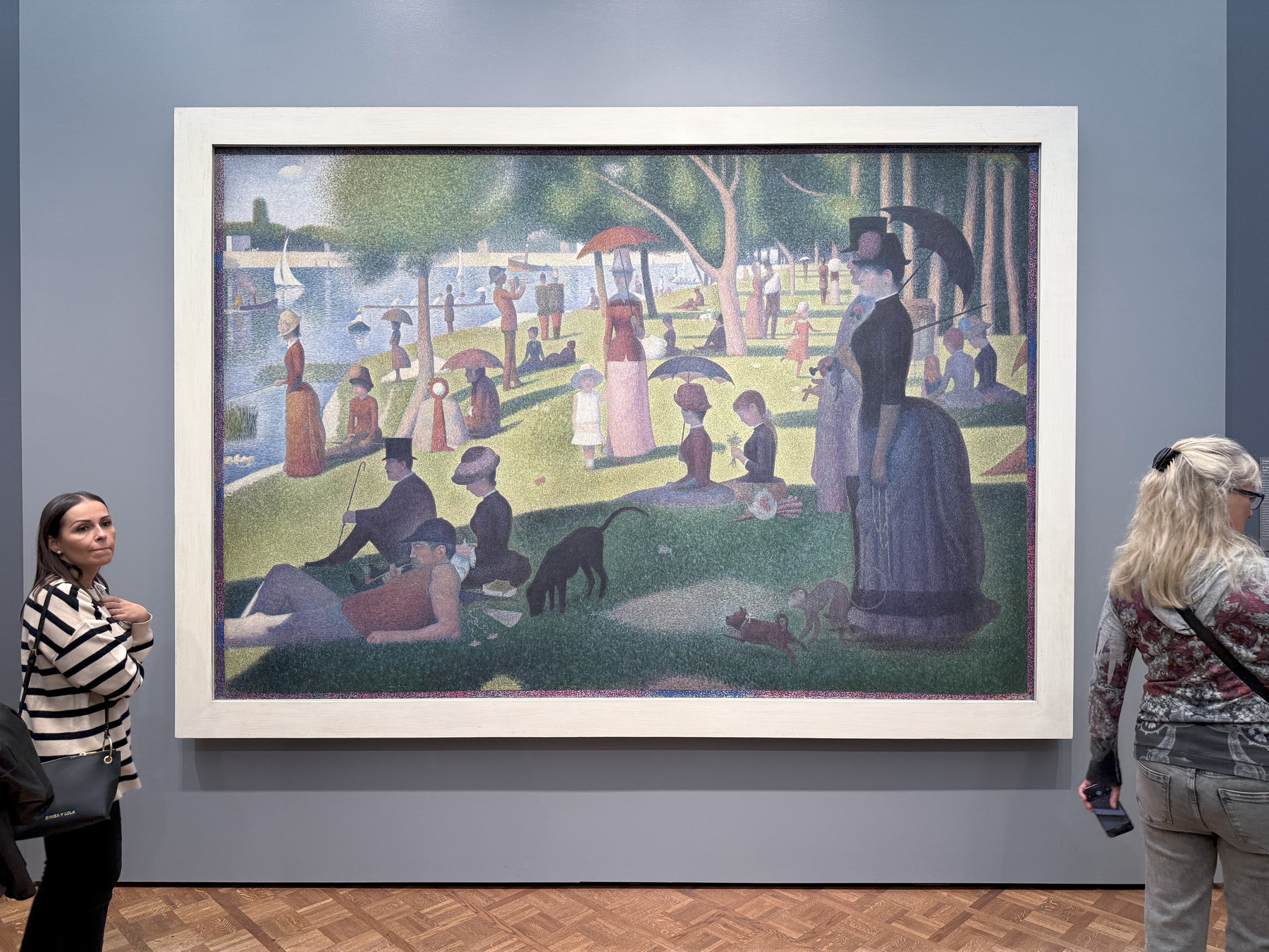 Auto-generated description: A large, framed pointillist painting depicting people relaxing in a park is displayed in a gallery, with two women observing it.