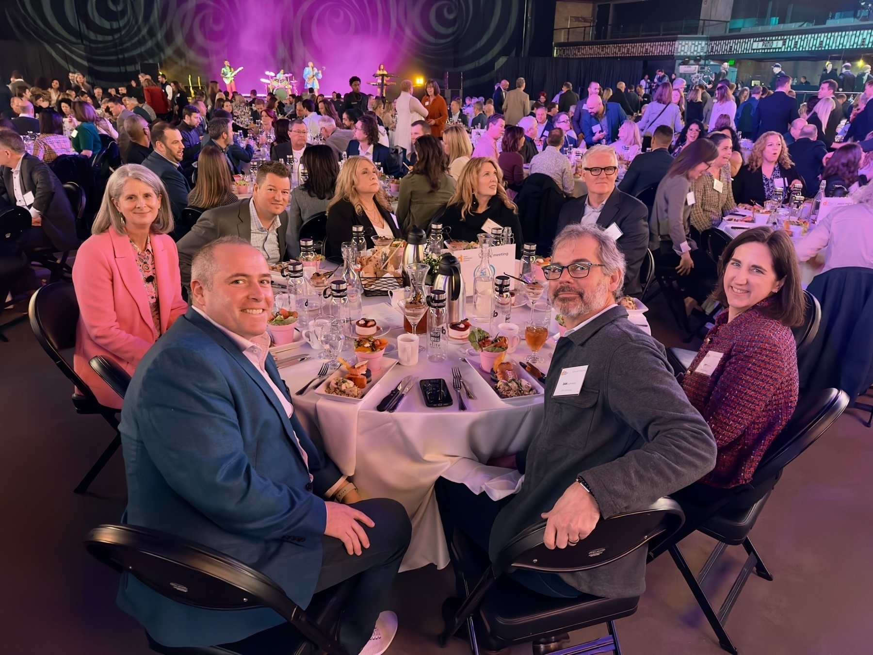 Leaders from SPS Commerce sitting at a dining table in a festive event hall with a live performance in the background.