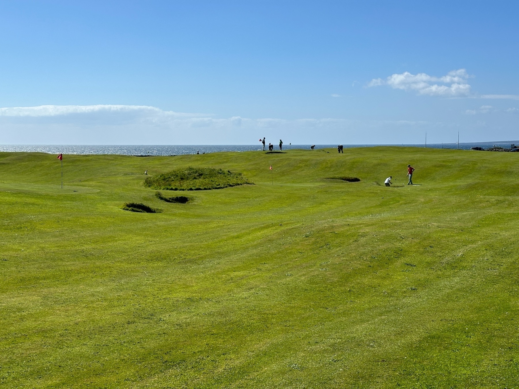 Auto-generated description: A grassy golf course near the ocean, with a few people in the distance under a clear blue sky.