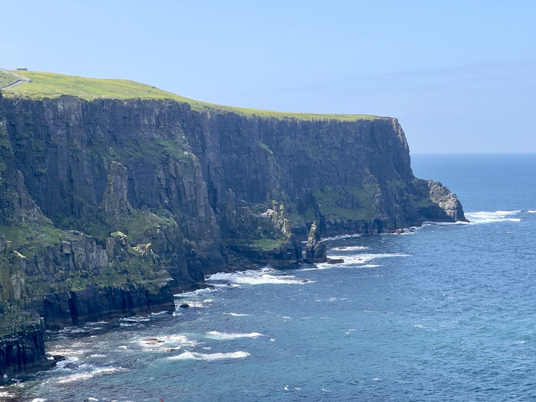 Auto-generated description: A dramatic cliffside meets the expansive ocean under a clear blue sky, with waves crashing against the rocky shore.