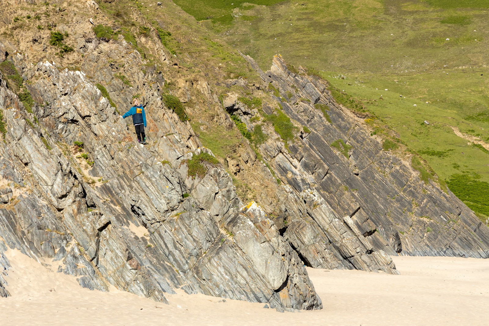 Auto-generated description: A person is climbing a steep, rocky slope on a coastal landscape with grass and sand.