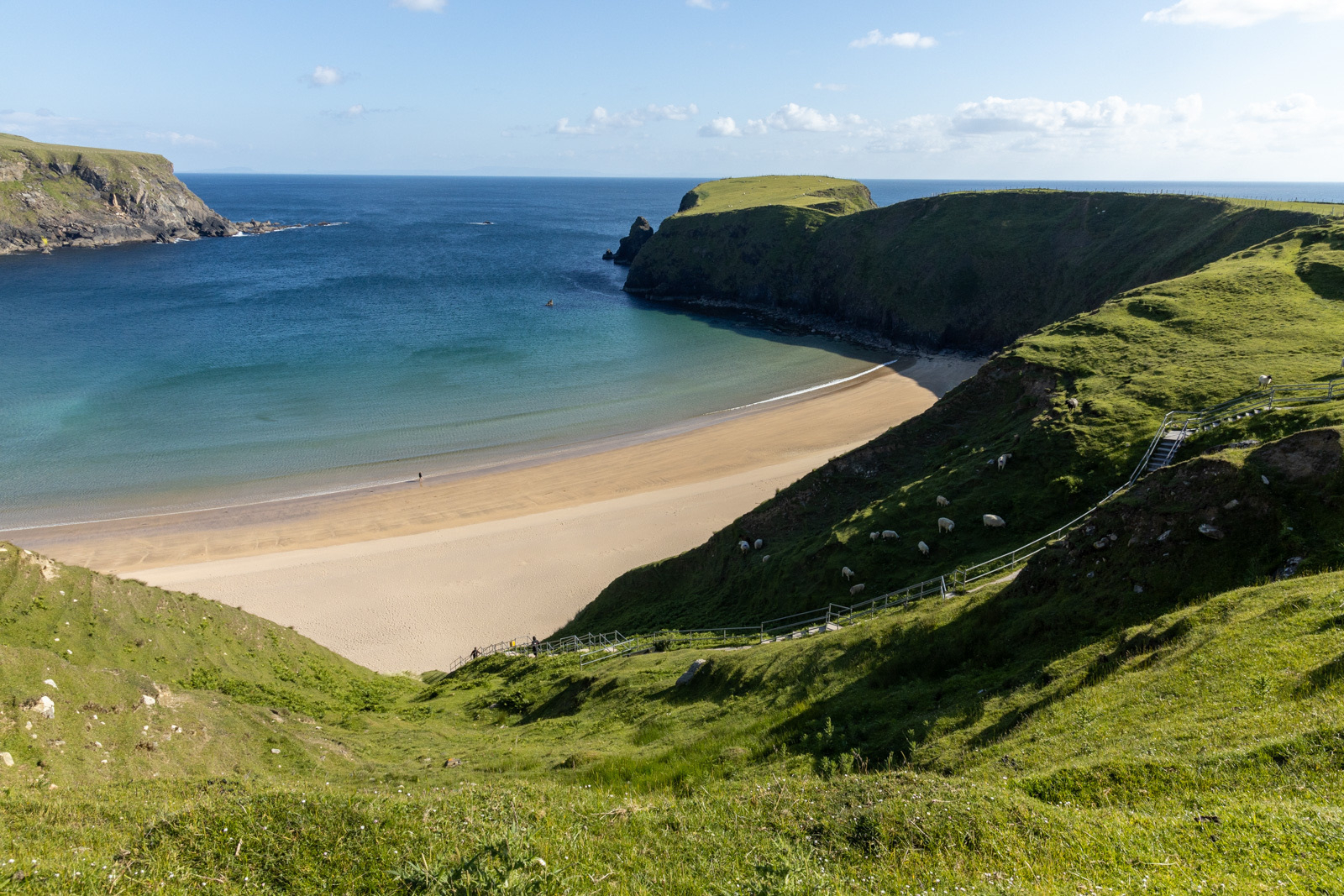 Auto-generated description: A serene coastal landscape features green cliffs surrounding a sandy beach, with the clear blue ocean extending into the horizon under a partly cloudy sky.