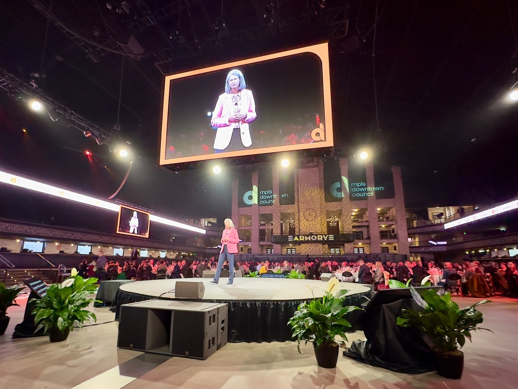 Karin Lucas on stage at a conference with a large audience, large screen display above showing the speaker, and banners indicating the event relates to the Minneapolis Downtown Council.