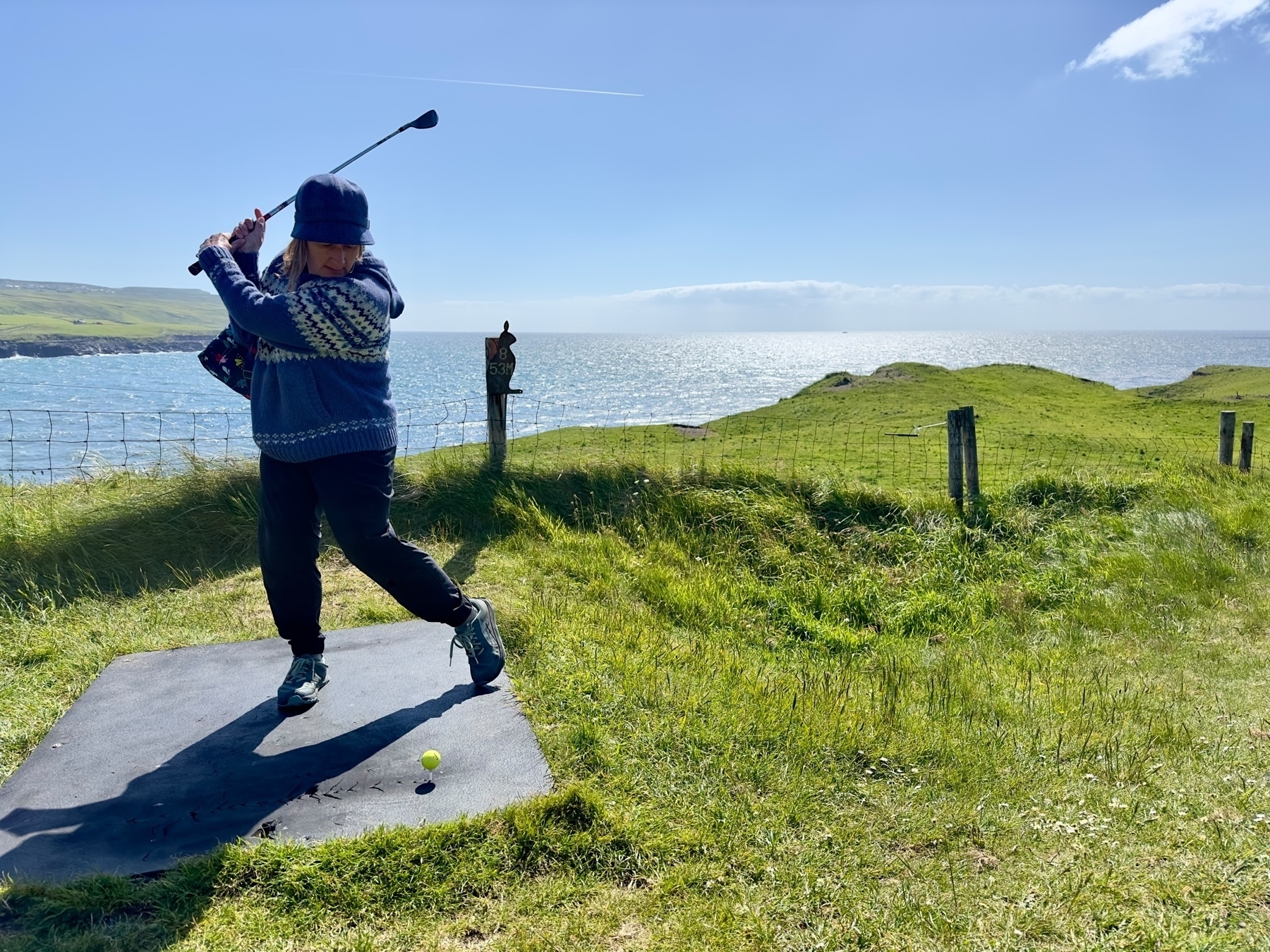 Auto-generated description: A person is swinging a golf club on a grassy coastal area with the ocean in the background.